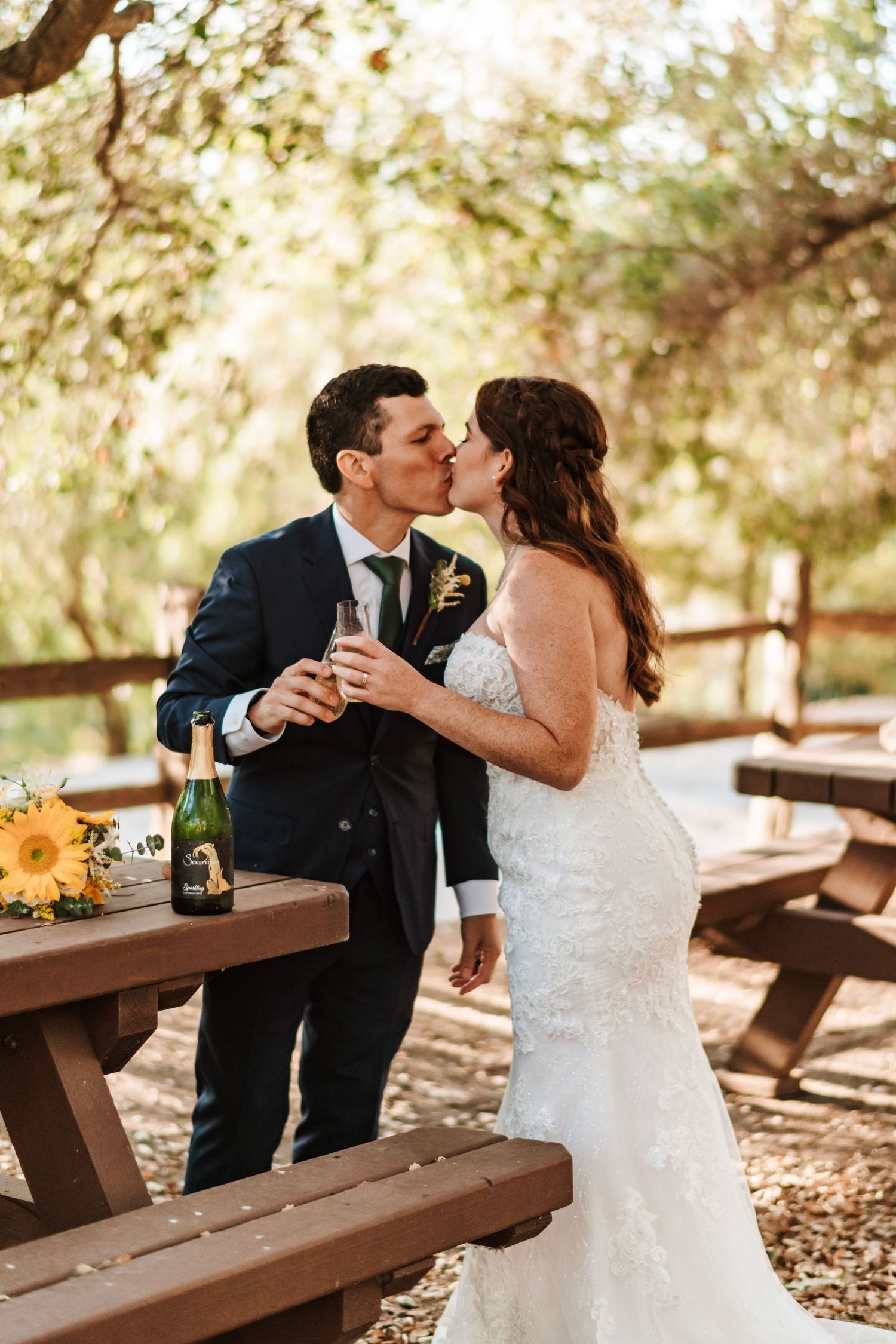 Bride and groom kiss beside picnic table with sunflowers and champagne outdoors.
