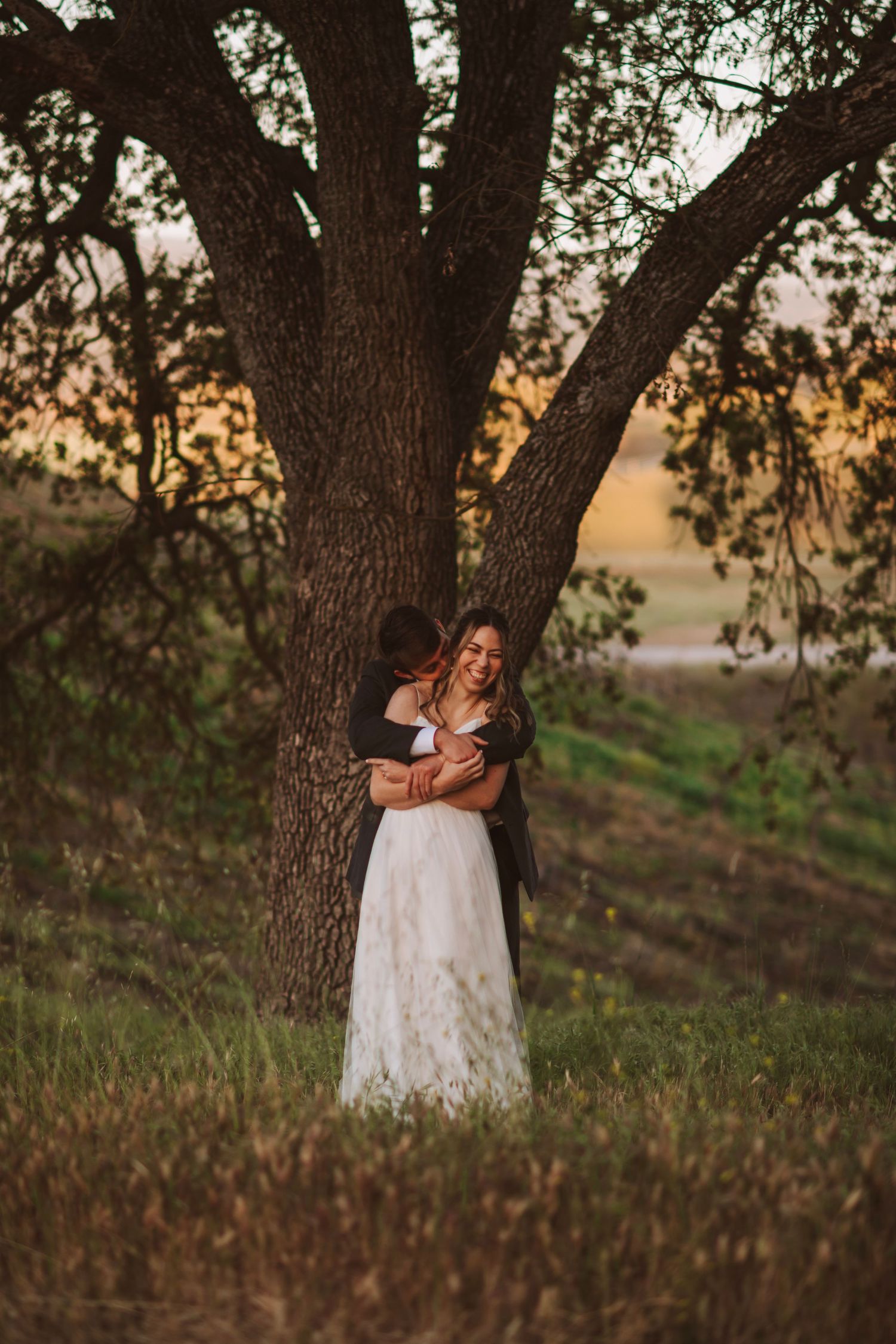 Groom embraces bride in white lace gown beneath a large oak tree at golden hour sunset.