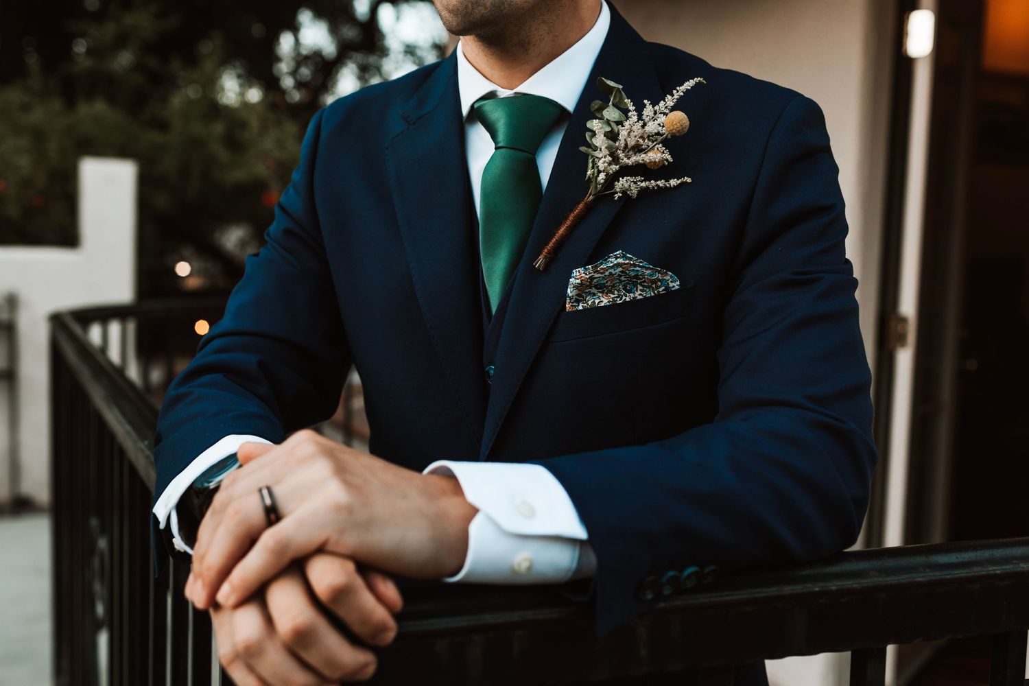 Close-up of groom in navy suit with green tie and floral boutonniere leaning on a railing.