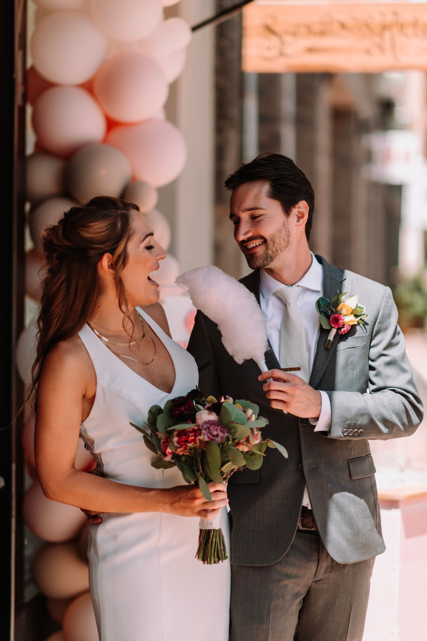 Bride and groom laugh while sharing pink cotton candy outside balloon-decorated venue.