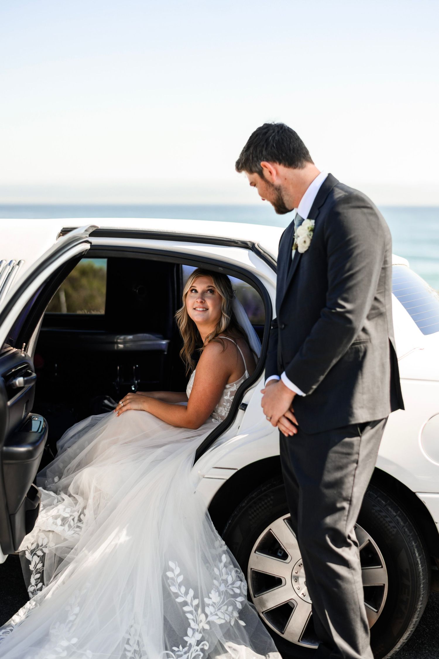 Smiling bride in lace gown sits in white limousine as groom in gray suit assists her by the sea.