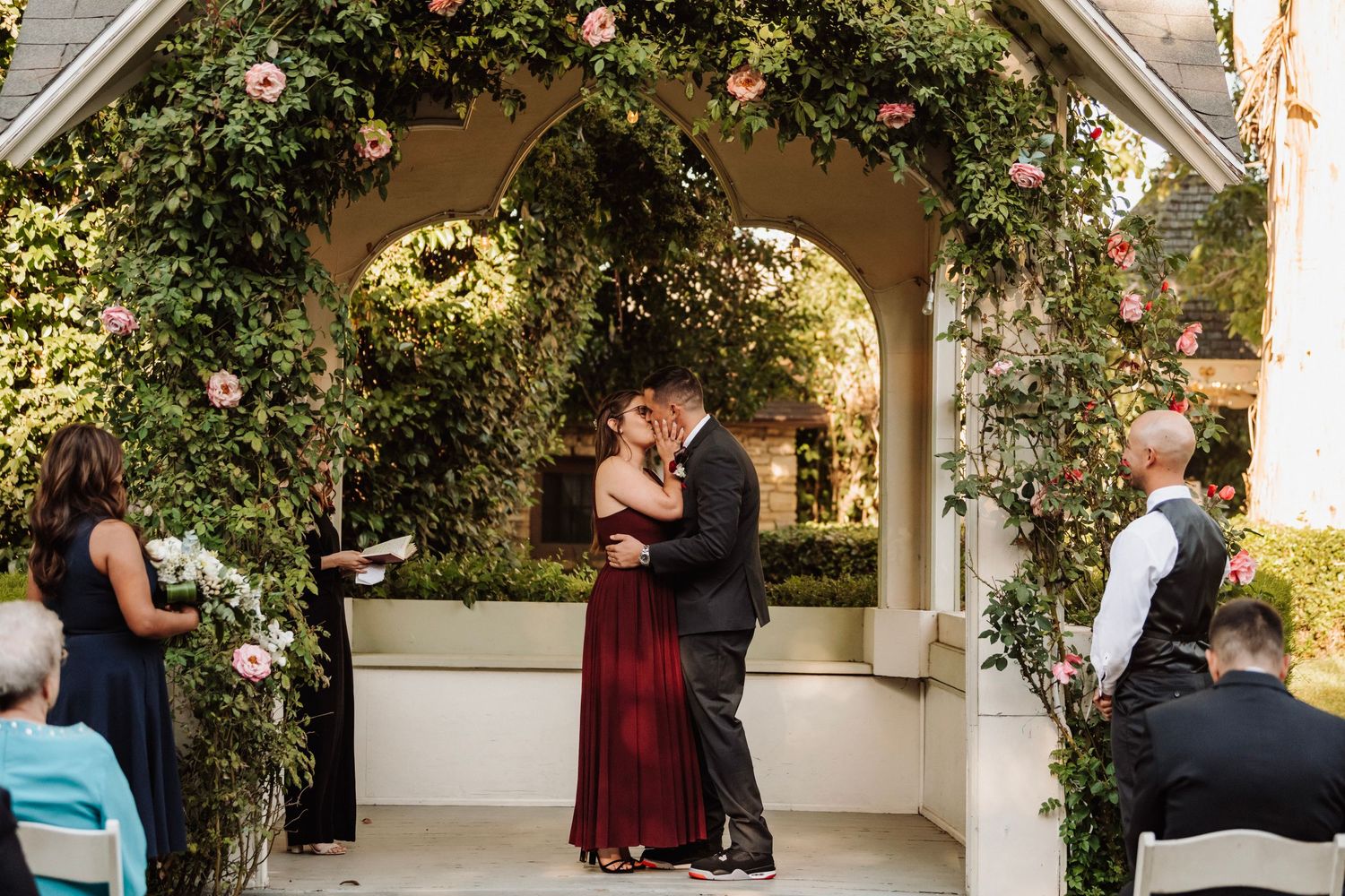 Couple shares first kiss under floral arch during outdoor wedding ceremony with guests watching.