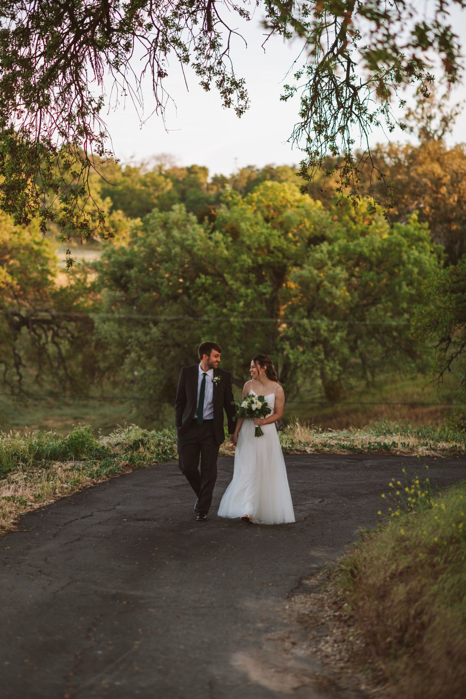 Bride and groom walk hand-in-hand down a tree-lined path surrounded by lush green foliage at sunset.