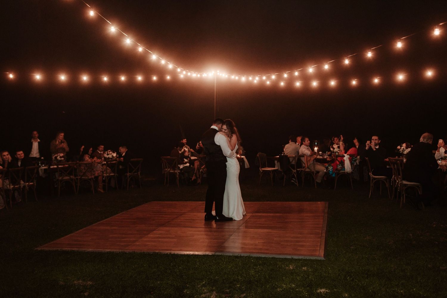 Couple shares first dance on wooden floor under string lights at night reception.