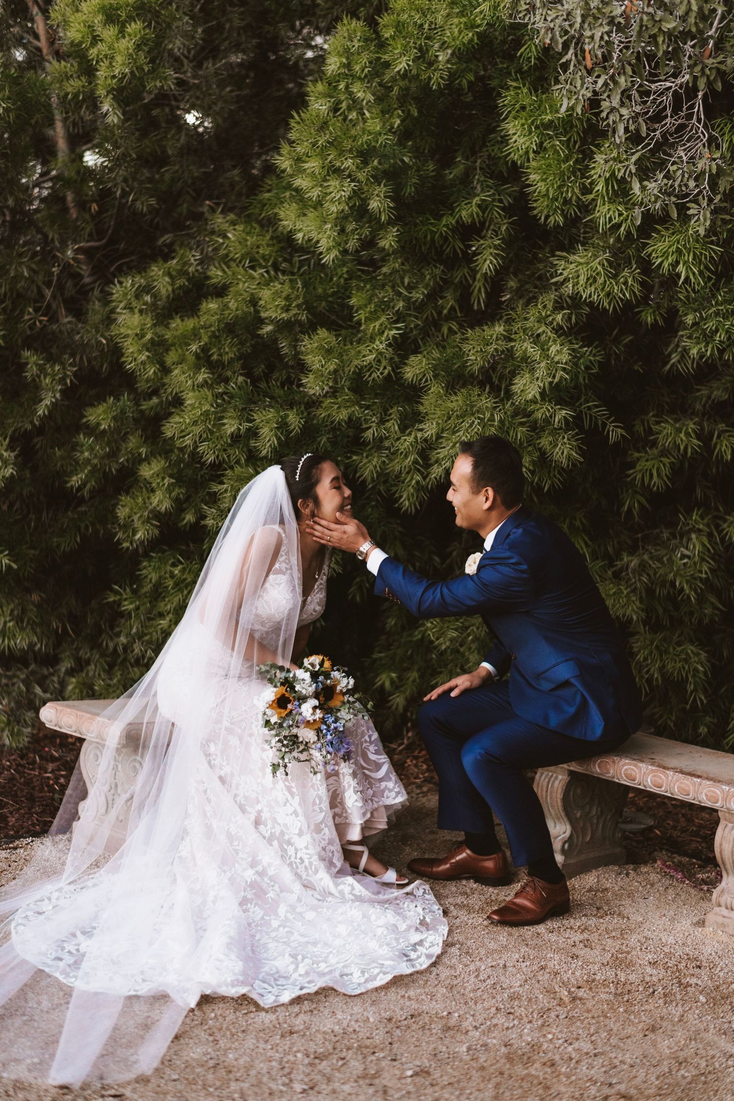 Groom in blue suit tenderly holds bride's face as they sit on stone bench surrounded by lush greenery.