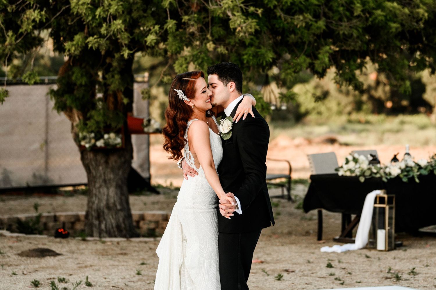 Newlyweds share a first kiss under a large tree at their outdoor wedding.