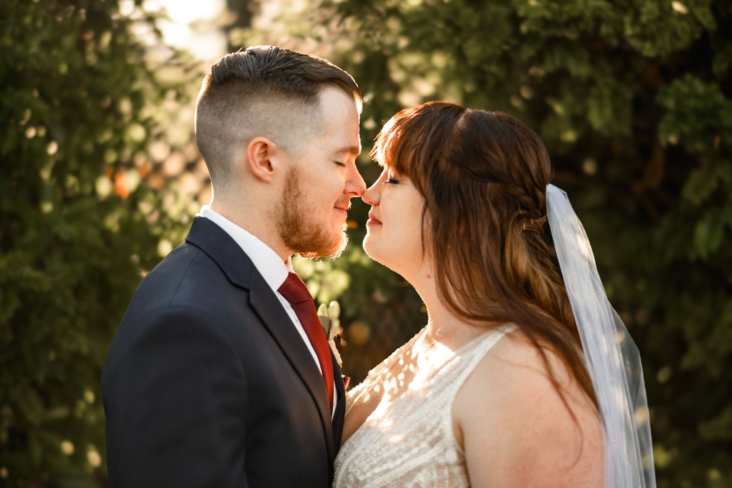Bride and groom sharing an intimate nose-to-nose moment in golden sunlight, bride wearing veil and lace gown.