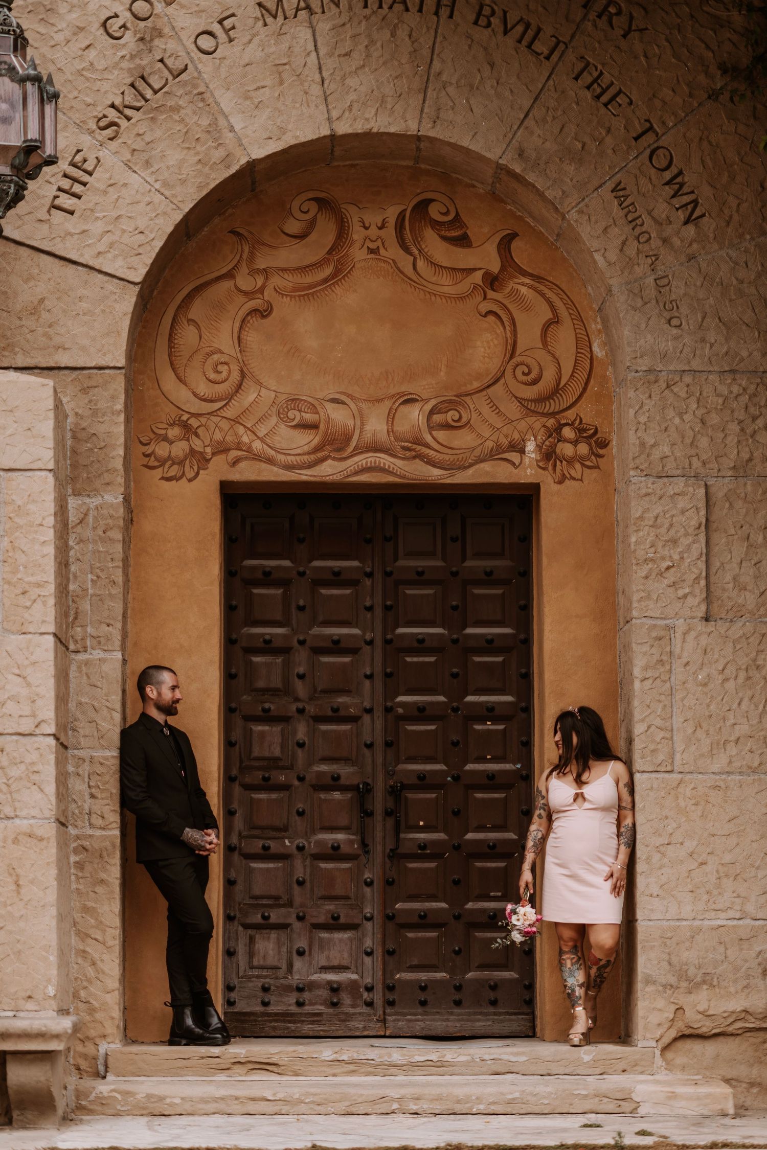 Bride and groom pose before ornate wooden doors of a historic stone building with arched entryway.