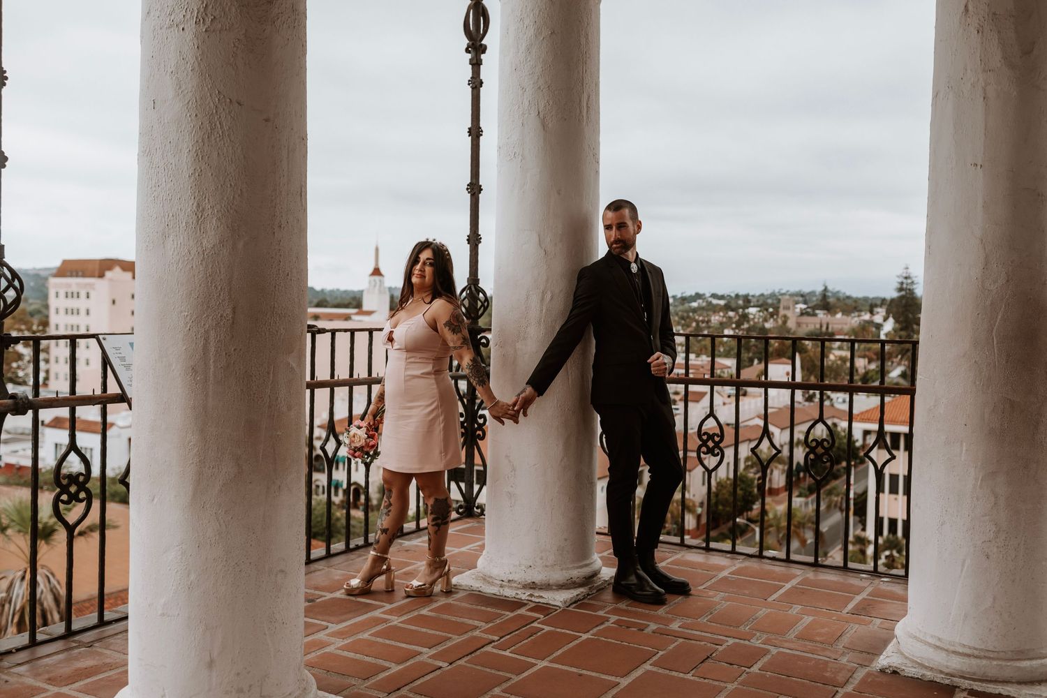 Couple holding hands on a terracotta balcony with white columns overlooking Santa Barbara cityscape.