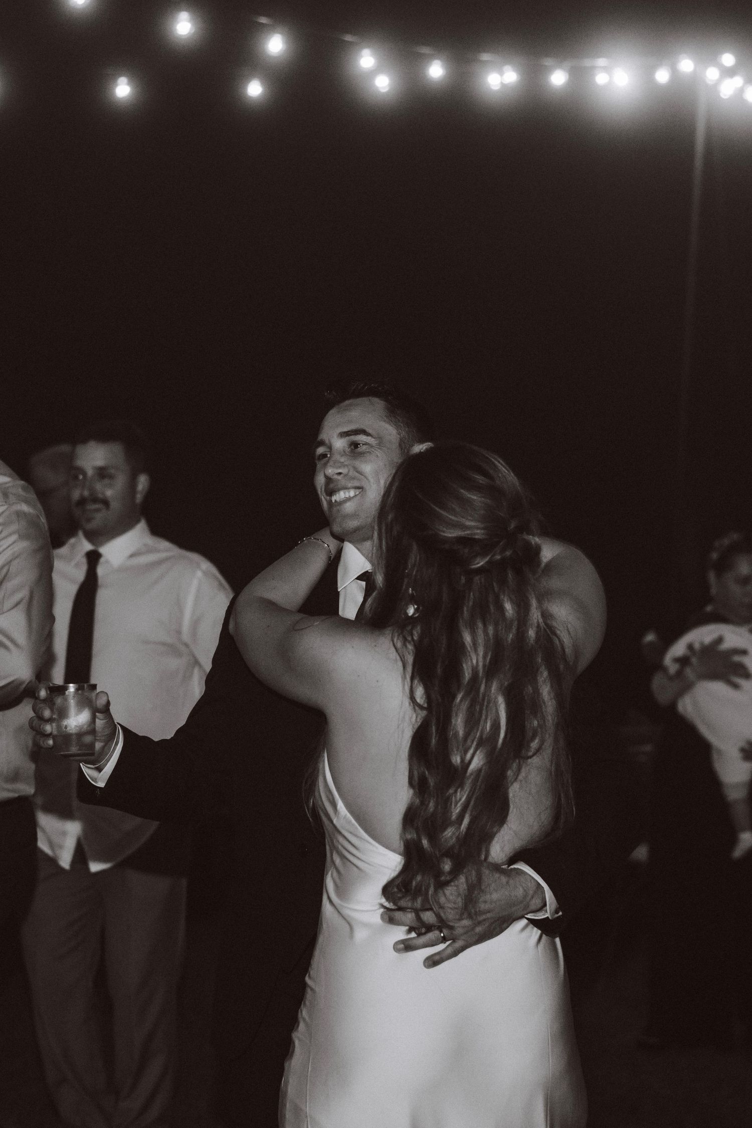 Black and white photo of smiling groom dancing with bride under string lights.