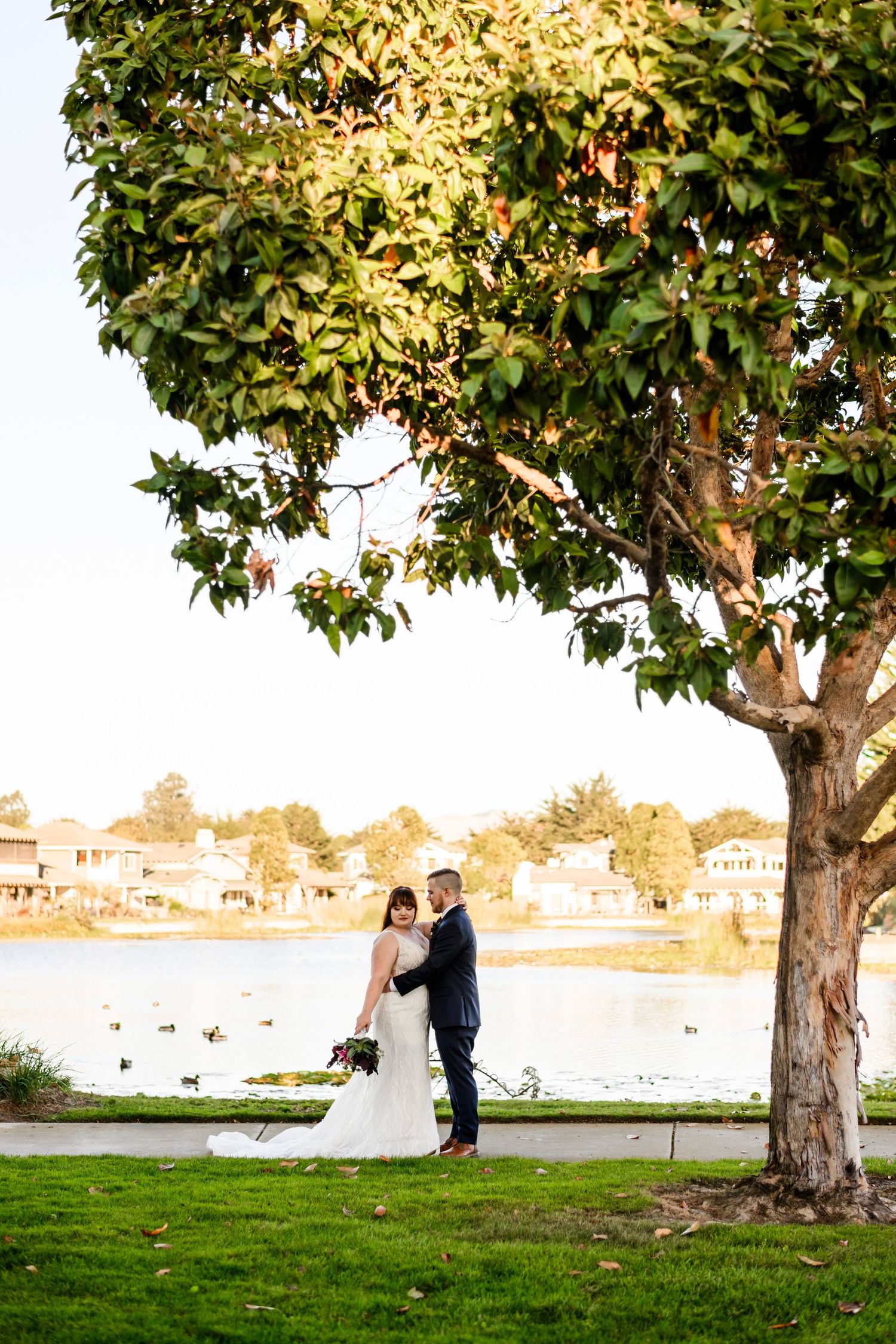 Bride and groom pose romantically under a tree by a serene lake at golden hour.