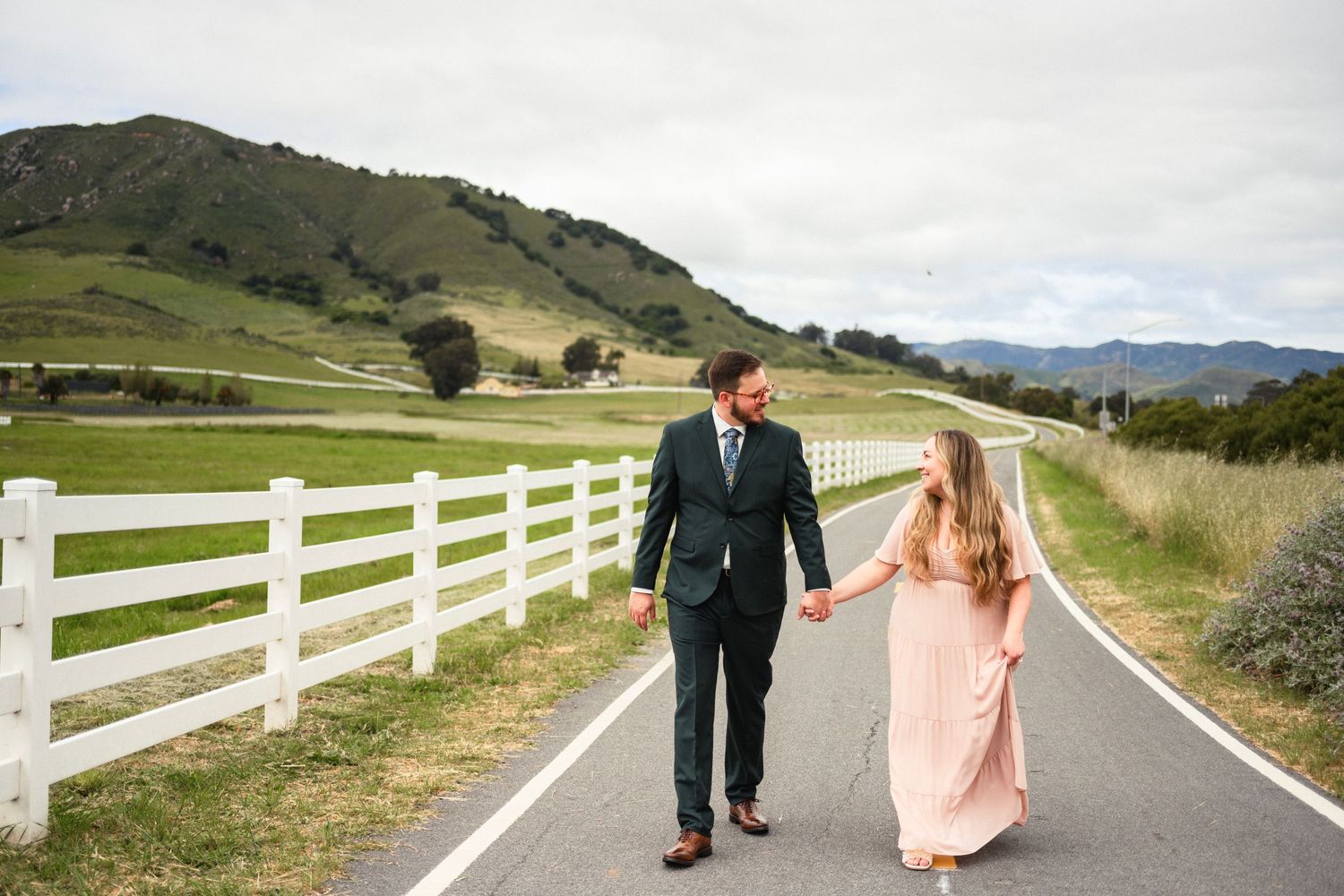 Couple walks hand in hand on rural road with white fence and rolling green hills in background.