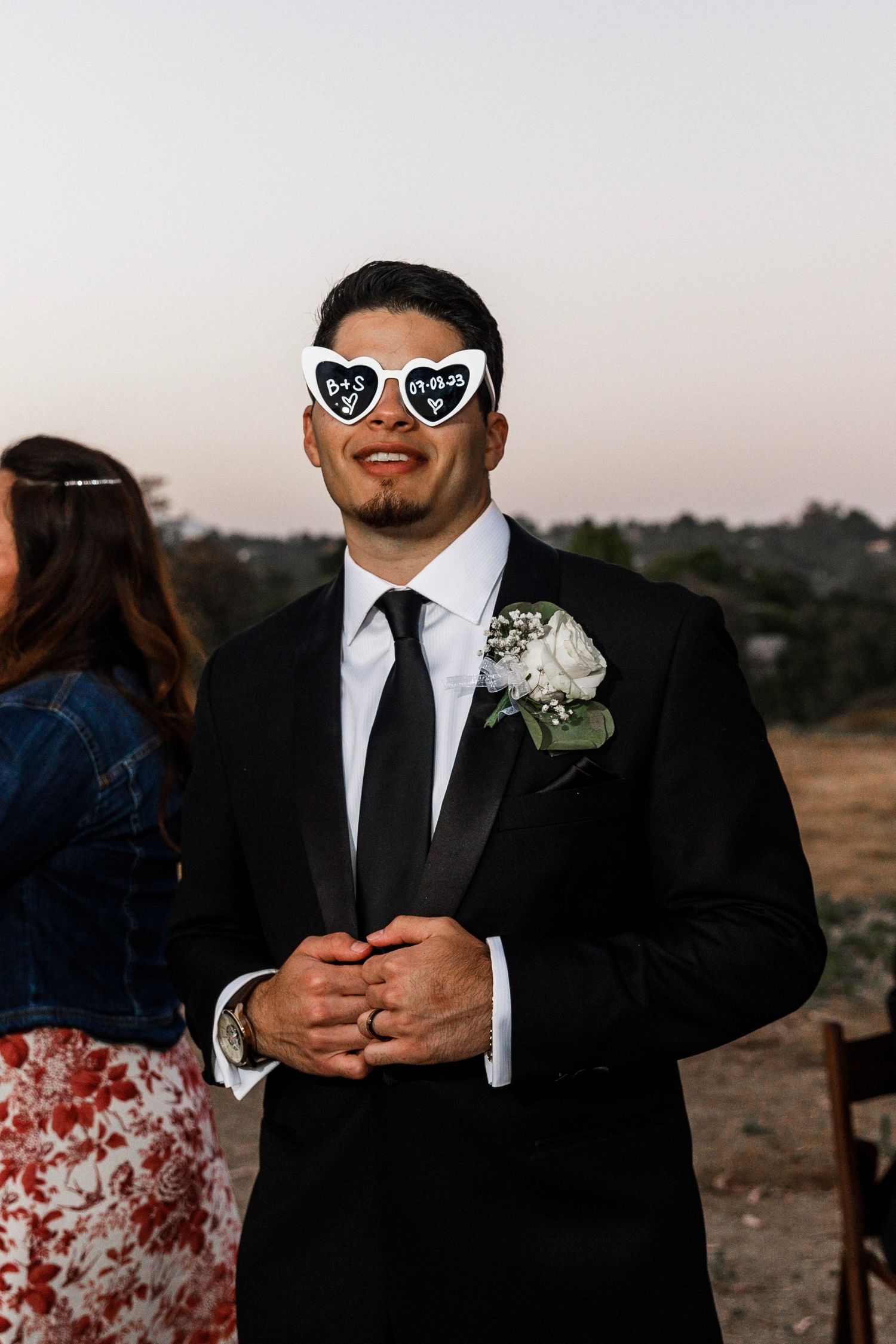 Groom in black tuxedo wearing novelty heart-shaped sunglasses with white rose boutonniere at outdoor wedding reception.