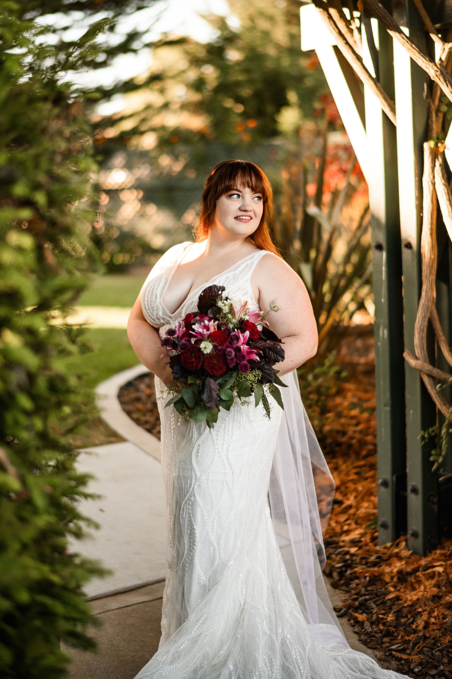 Bride in white lace gown holding a deep red bouquet standing in a sunlit garden archway.