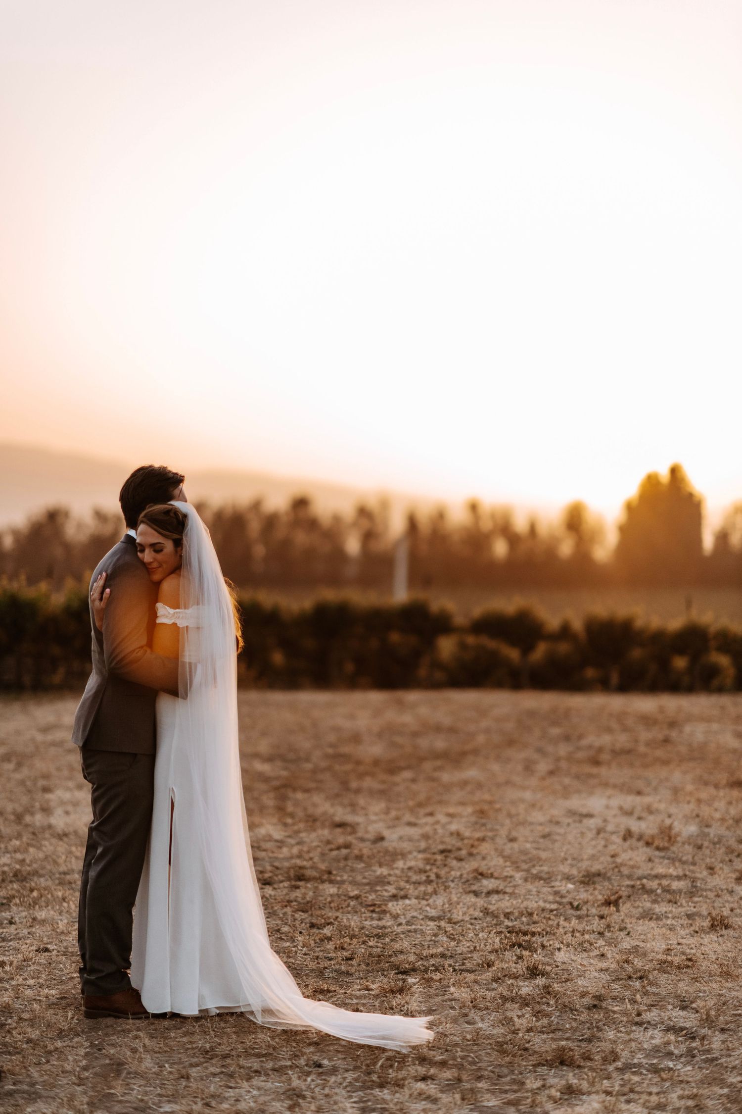 Bride and groom embrace tenderly in golden sunset light in an open field.