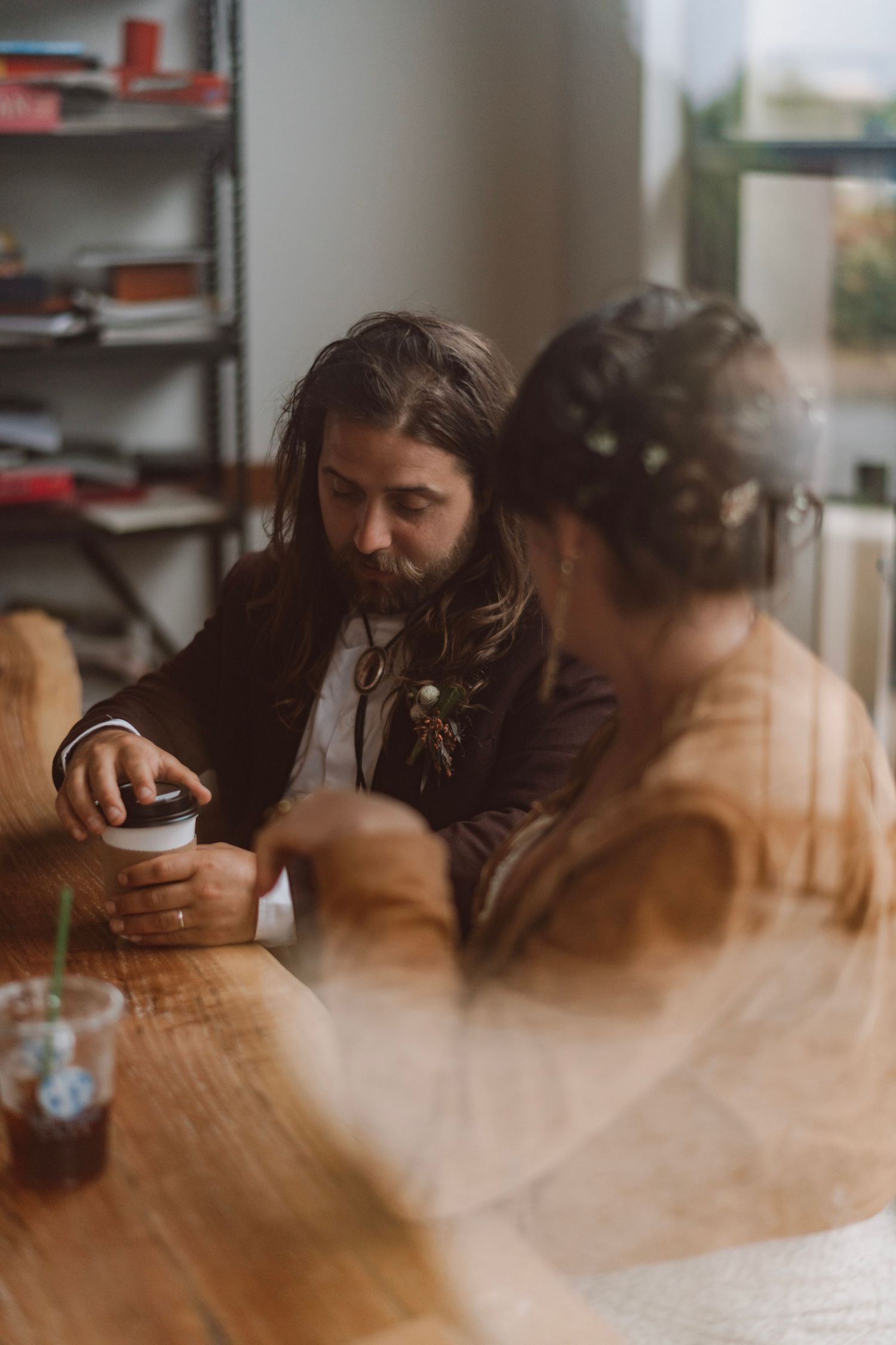 Long-haired groom and bride sit together at a wooden table in a warmly lit cafe setting.