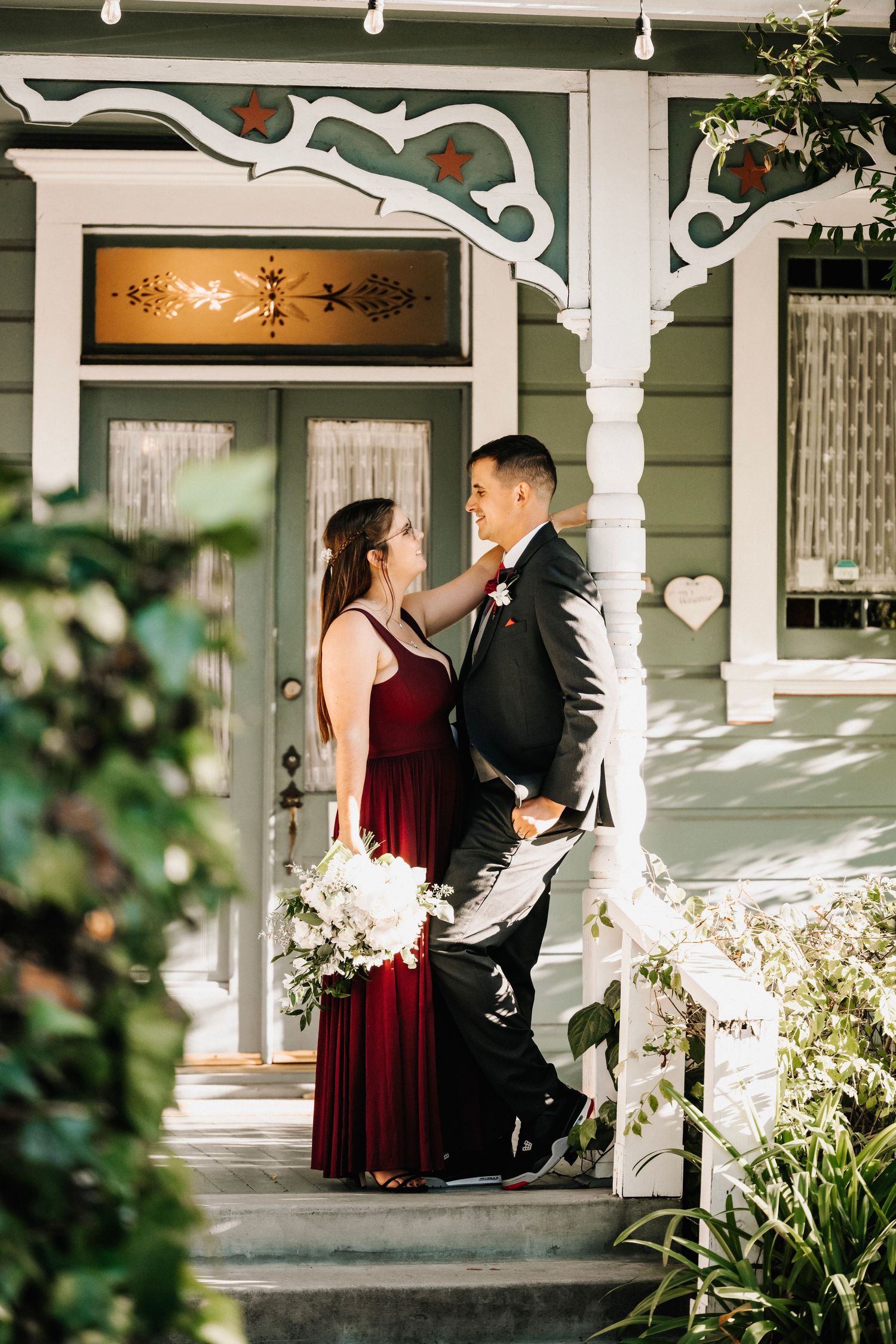 Bride in red velvet gown and groom in black suit share romantic moment on vintage home porch.
