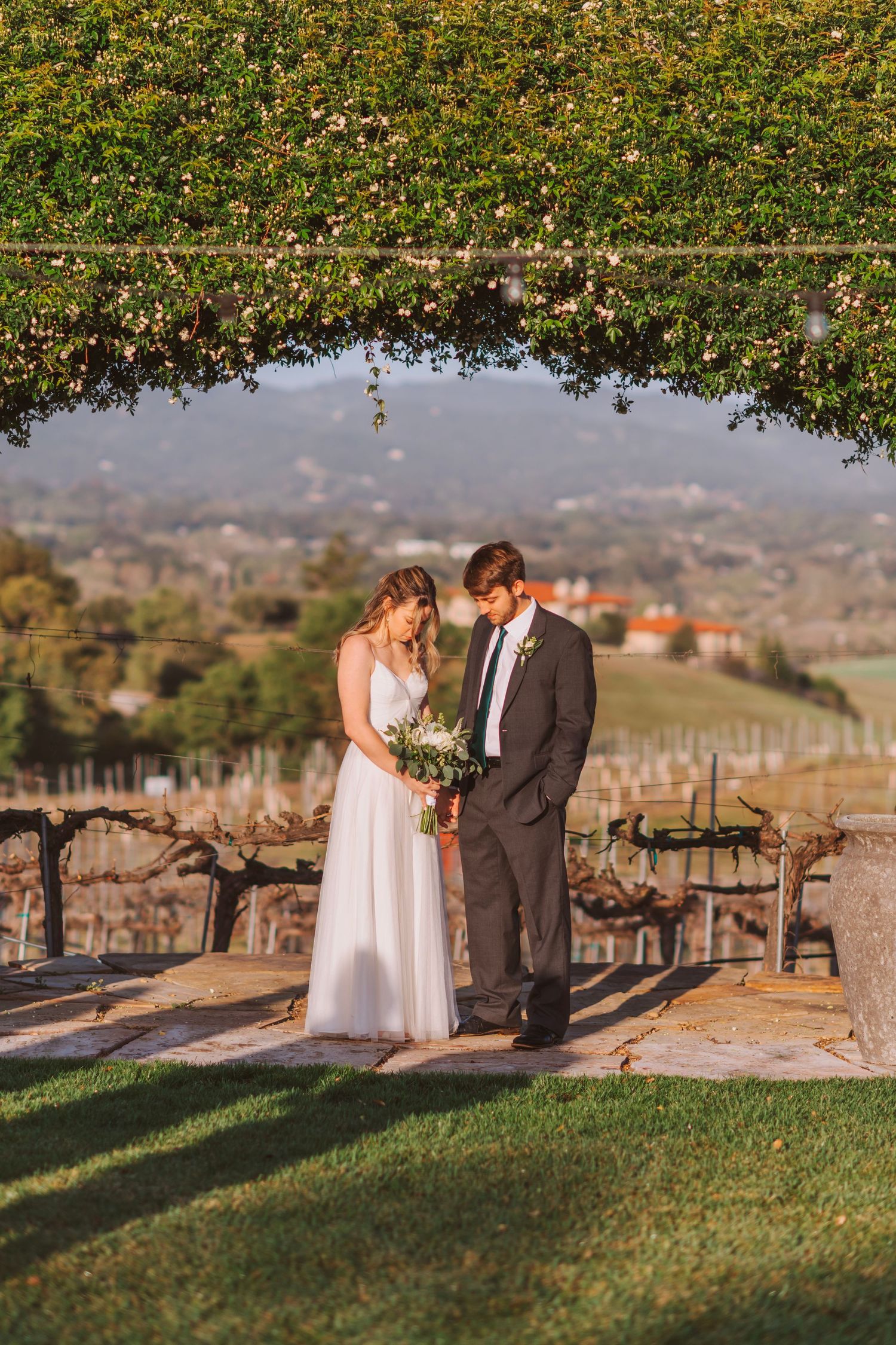 Bride and groom admire bouquet on stone terrace with scenic vineyard valley views at golden hour.