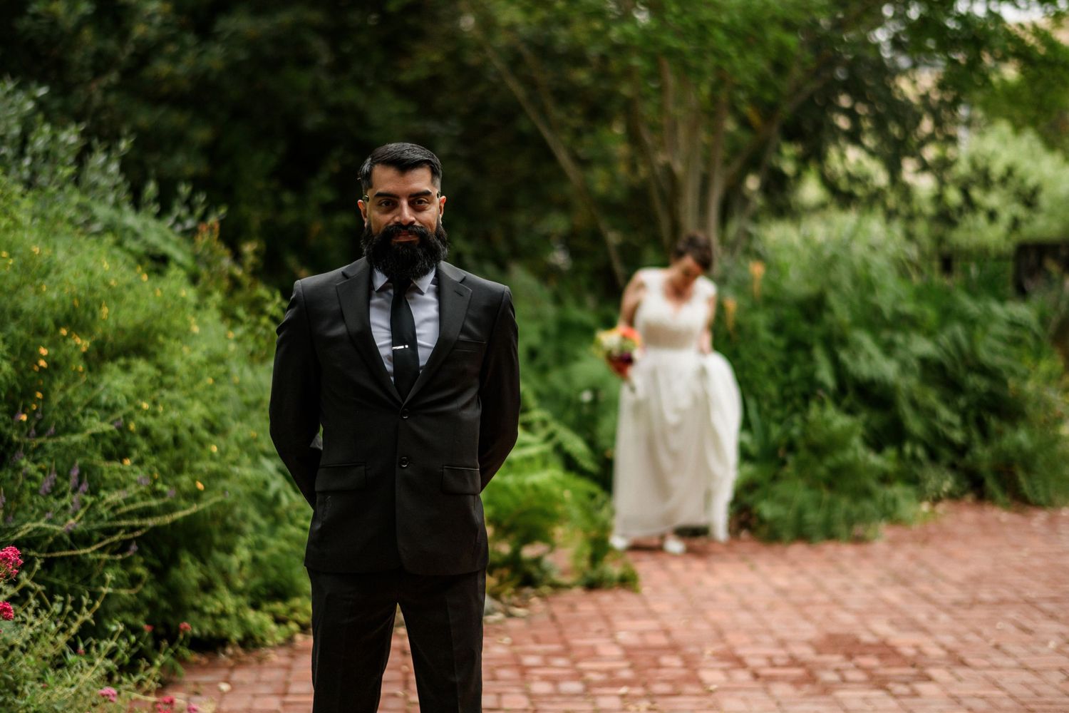 Groom in black suit stands on brick path in garden while bride in white gown approaches in background.