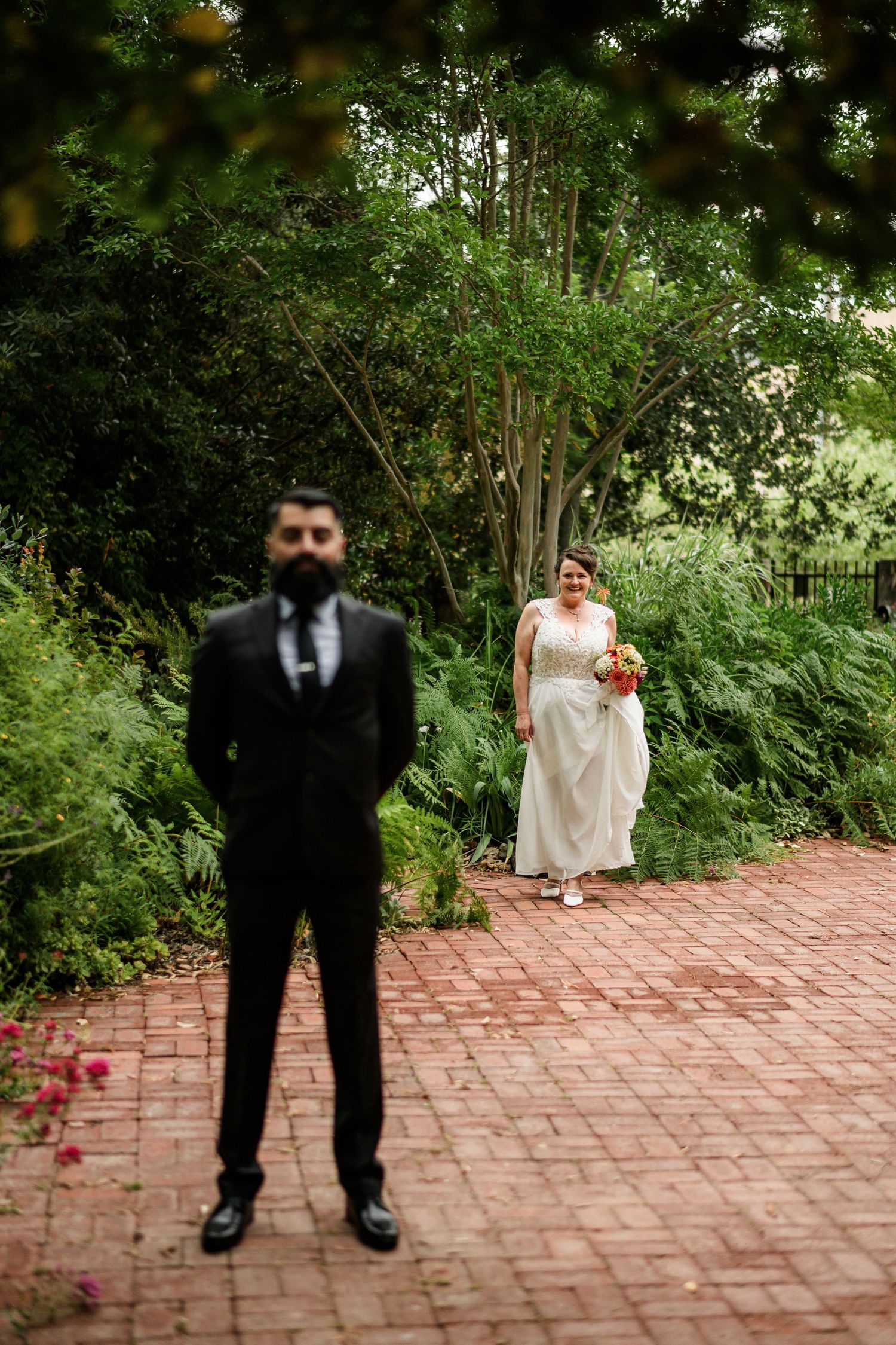 Groom in black suit stands in foreground on brick path as bride carrying bouquet walks toward him in lush garden.