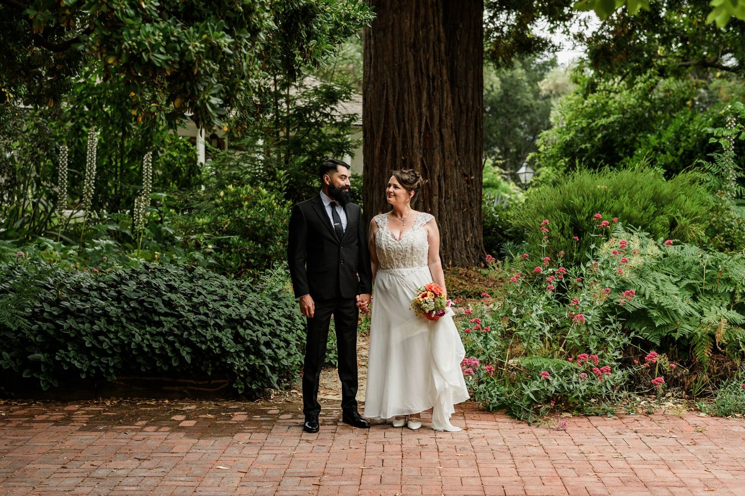 Bride and groom stand together on brick path holding hands near large redwood tree in lush garden setting.