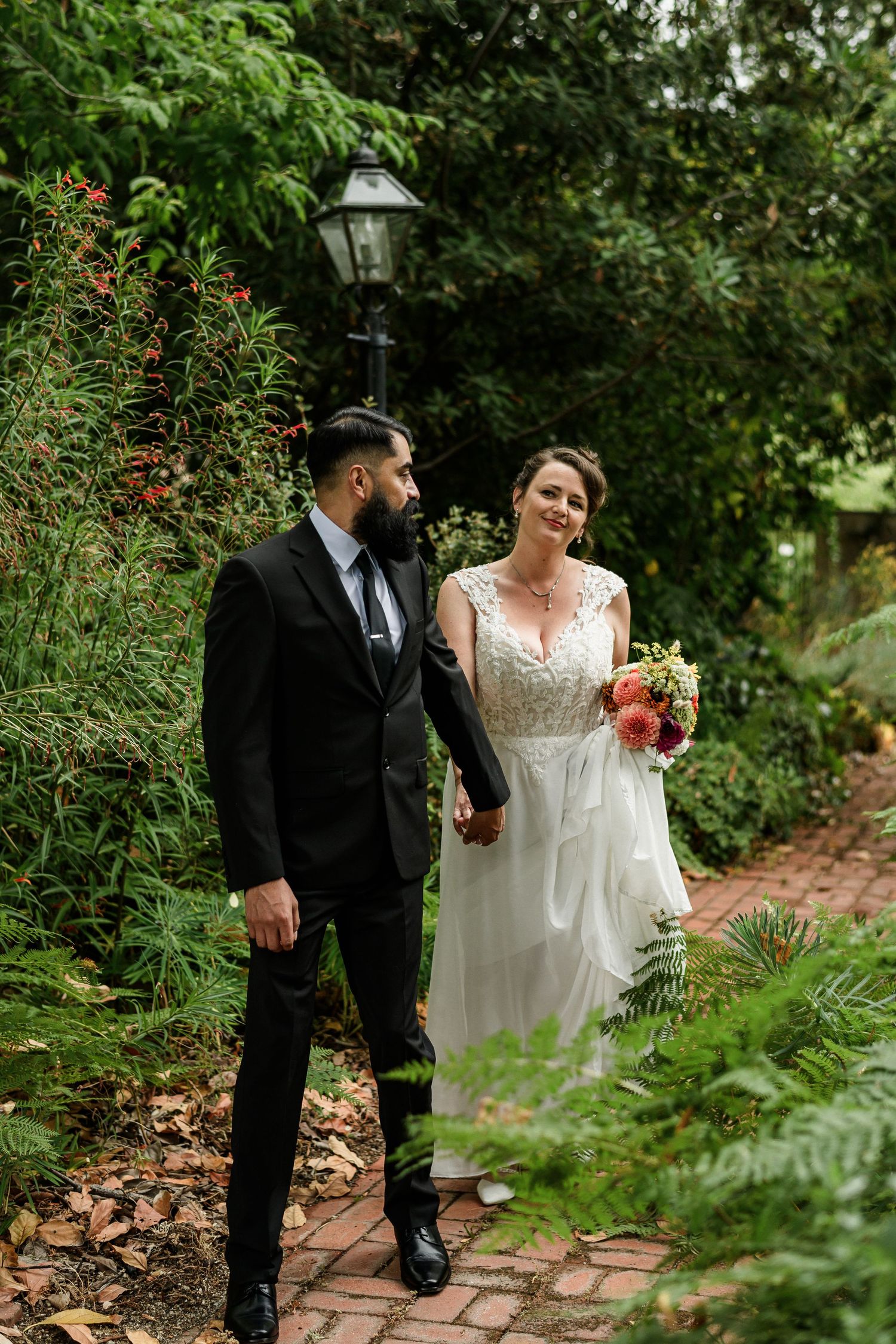 Bride in white lace gown holds bouquet while walking with bearded groom in black suit through a lush garden path.