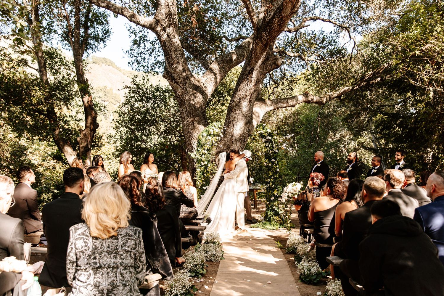 Outdoor wedding ceremony under a large oak tree with guests seated along a garden aisle.