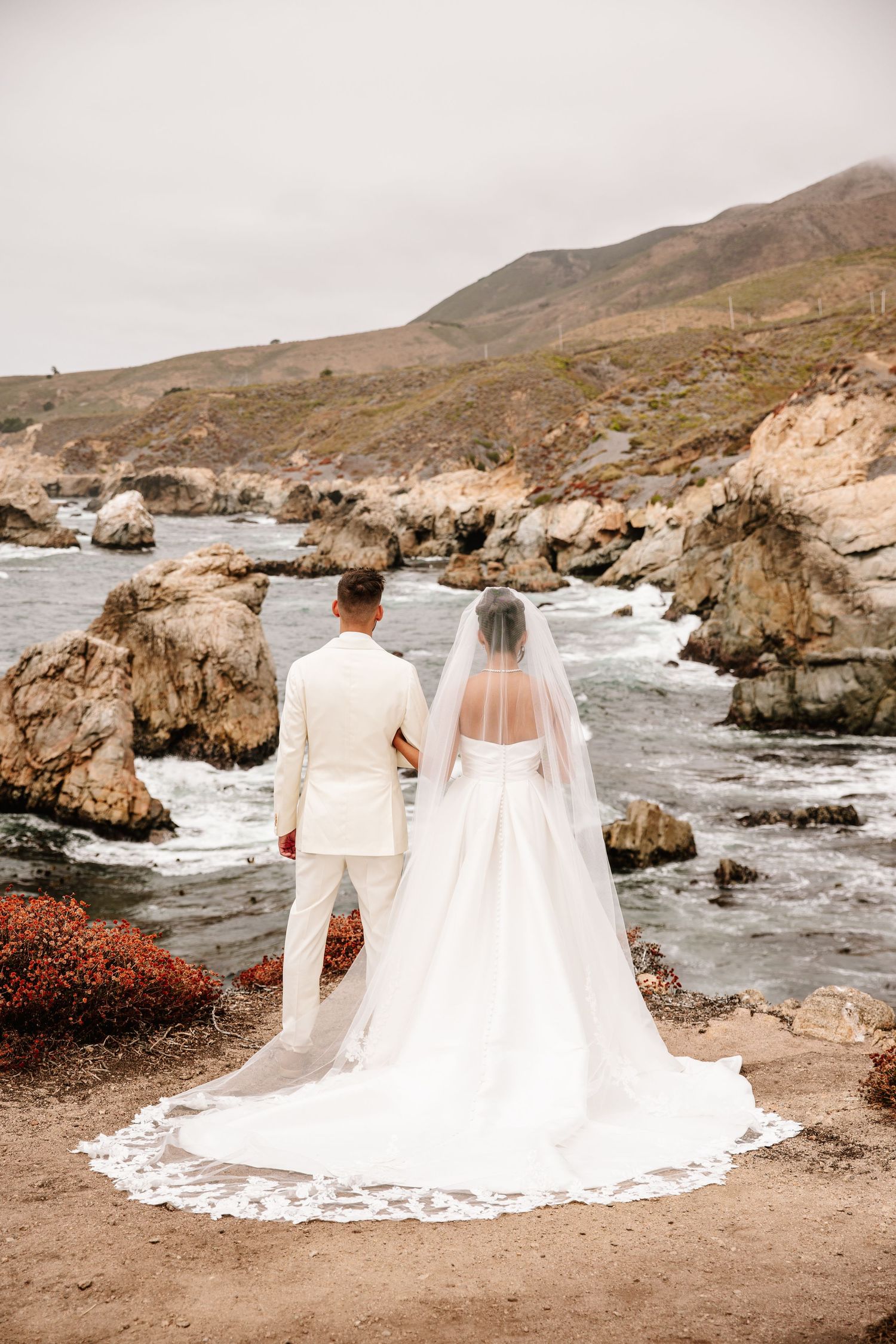 Bride and groom stand hand in hand overlooking a dramatic rocky coastal shoreline.