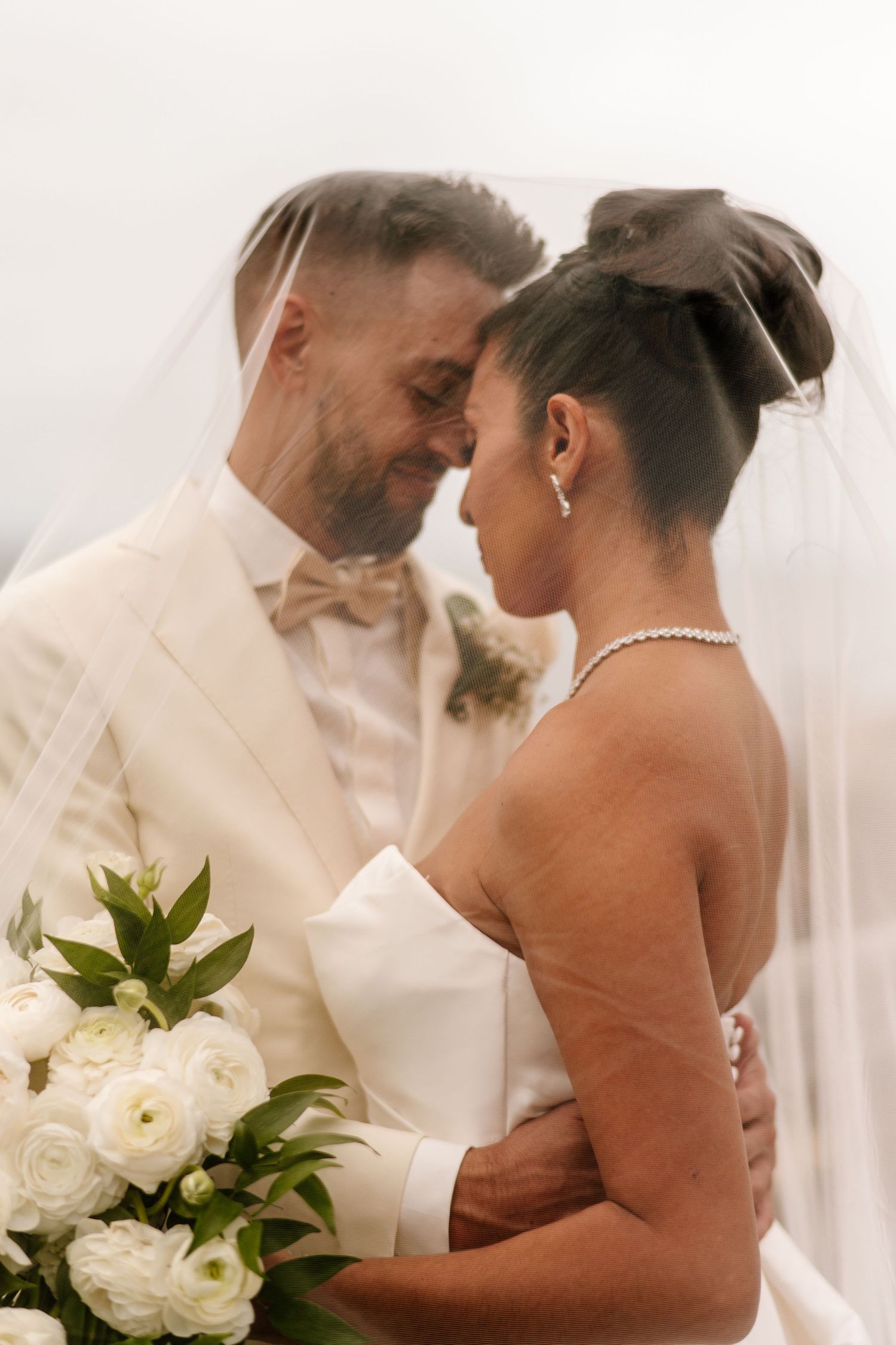 Bride and groom share an intimate moment under a flowing veil with white floral bouquet.