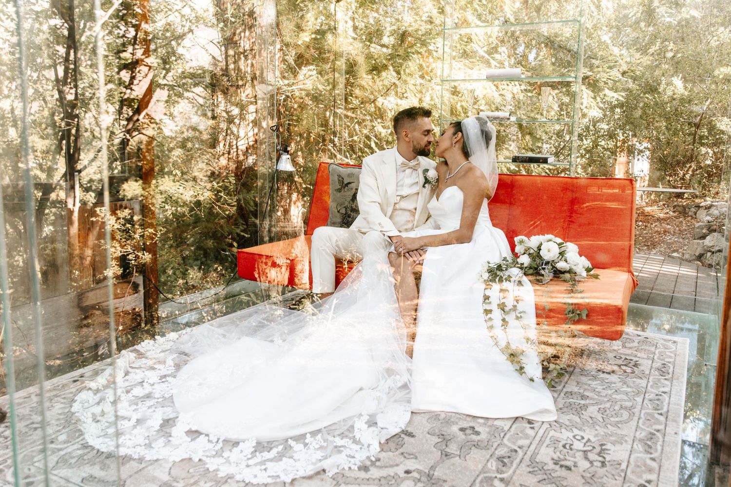Bride and groom share a kiss on a red bench inside a glass greenhouse.