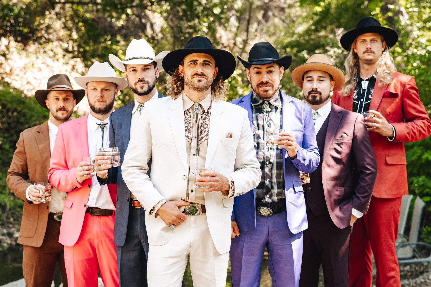 Group of seven groomsmen in colorful suits and hats posing outdoors.