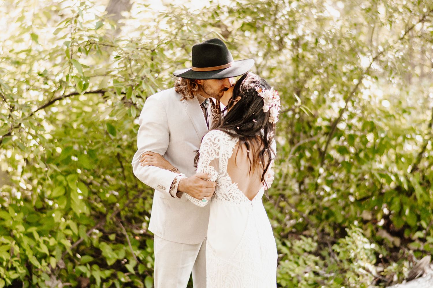 Bohemian bride and groom sharing a kiss surrounded by lush green foliage.