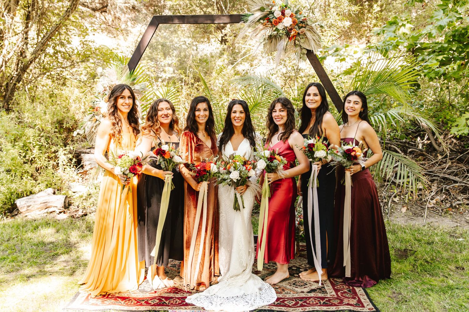 Bride with six bridesmaids in mismatched colorful dresses under a hexagonal arch.