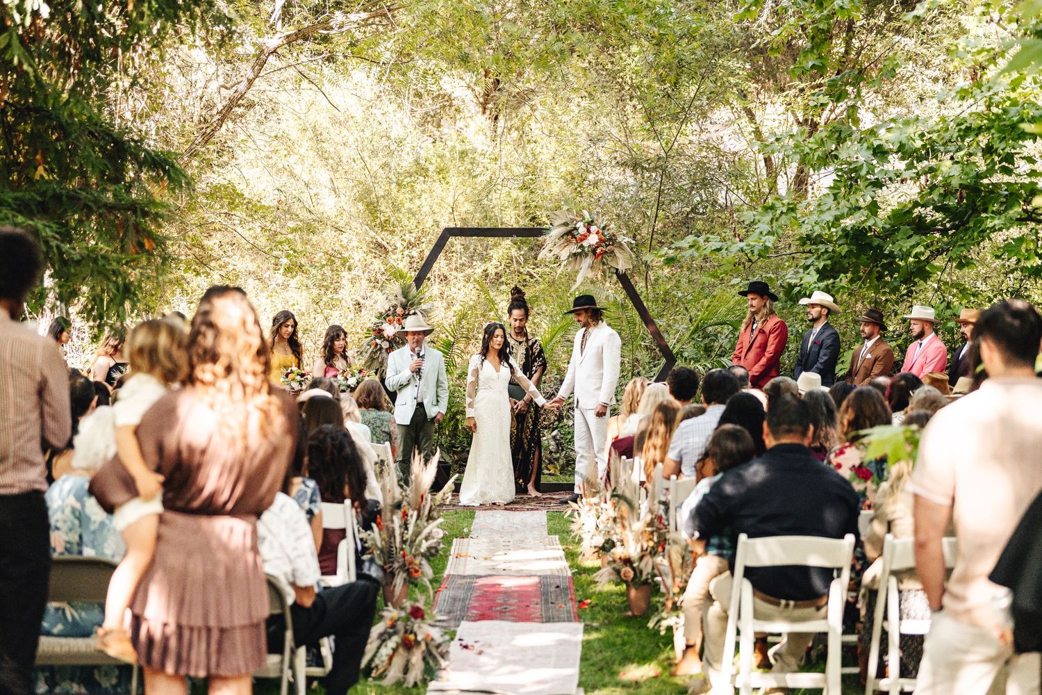 Outdoor boho wedding ceremony with guests seated along a decorated aisle in a garden.