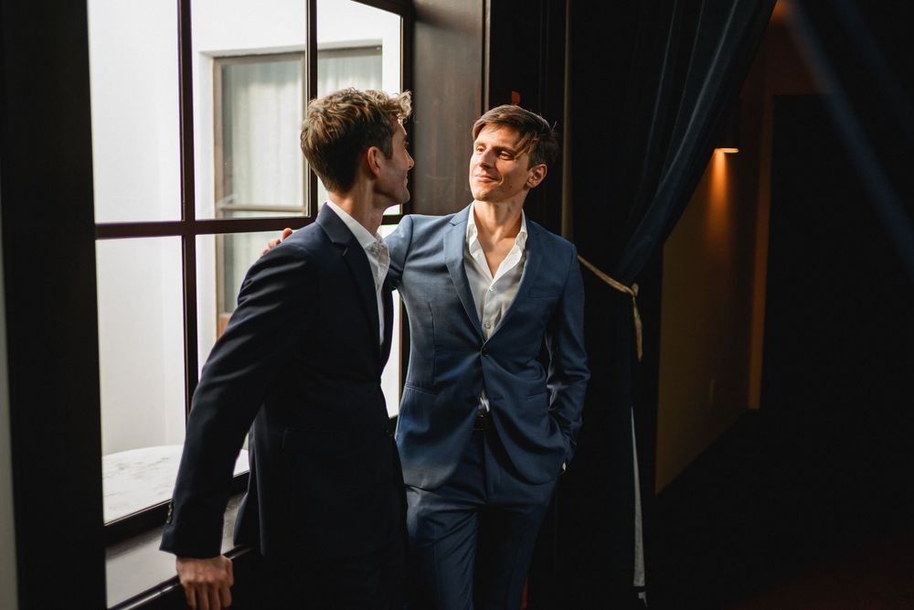 Two men in navy suits sharing an intimate moment by a window in elegant interior.