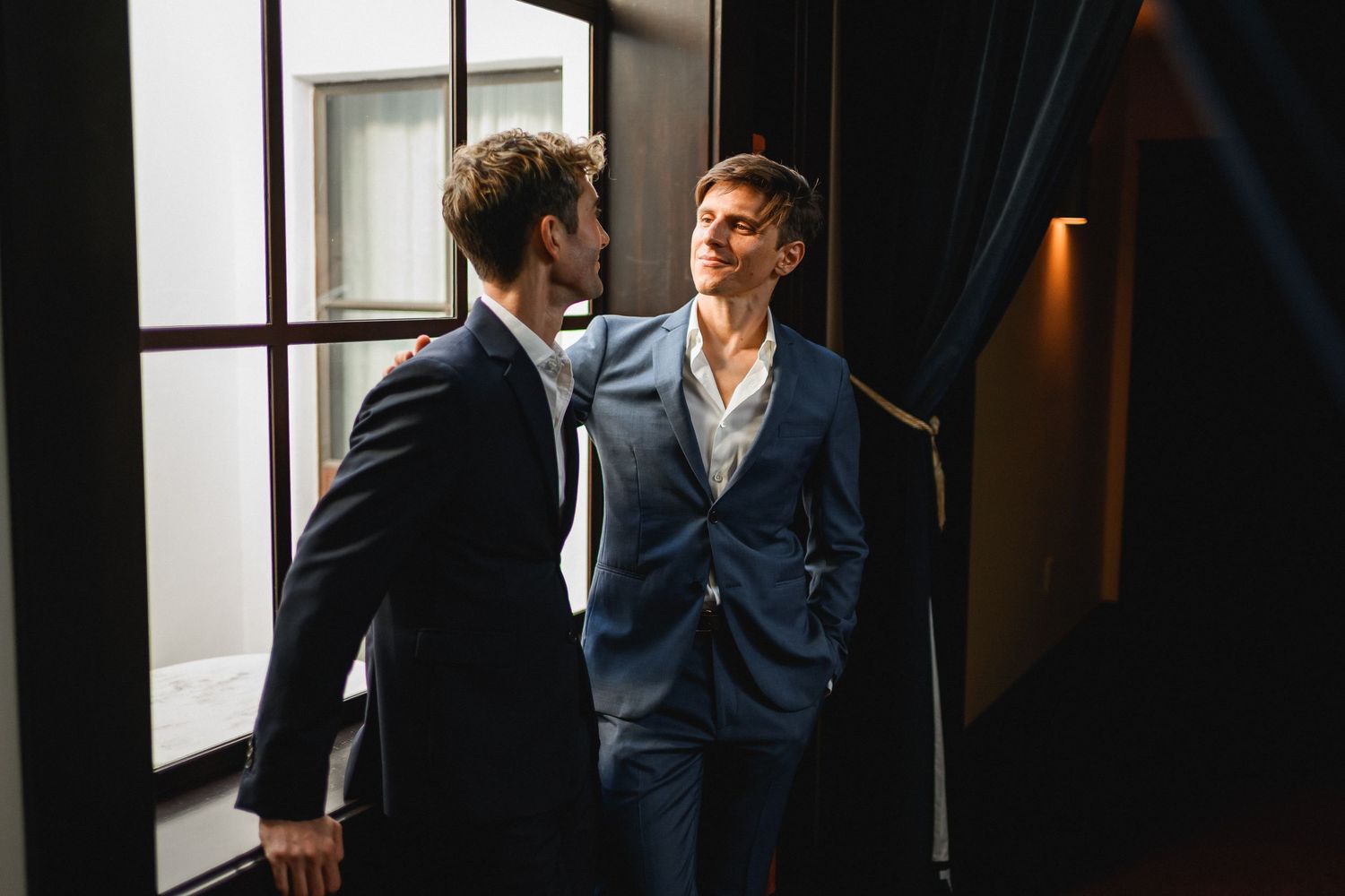Two men in navy suits sharing an intimate moment by a window in elegant interior.