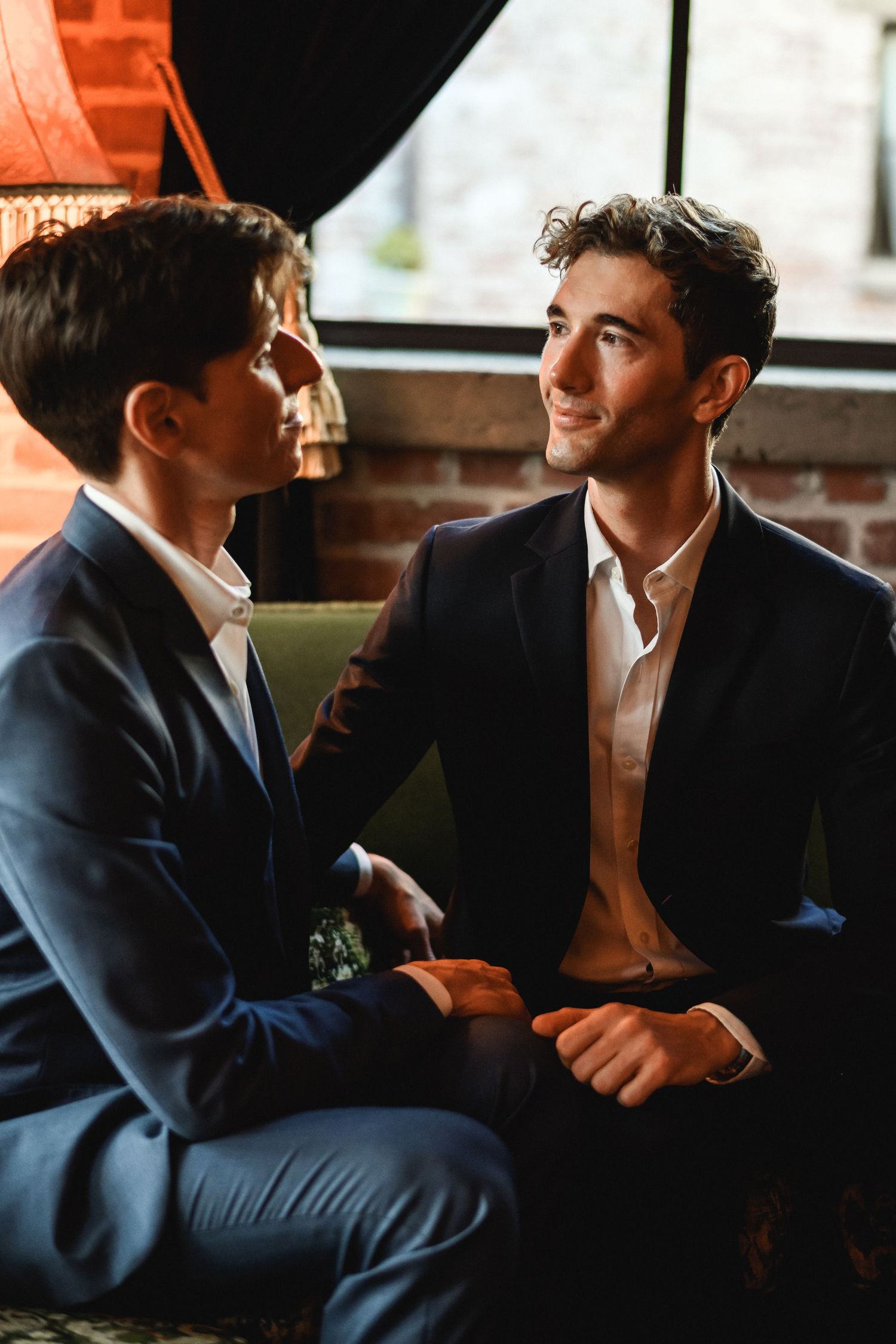 Two grooms in navy suits sitting together, smiling and holding hands in a cozy setting.
