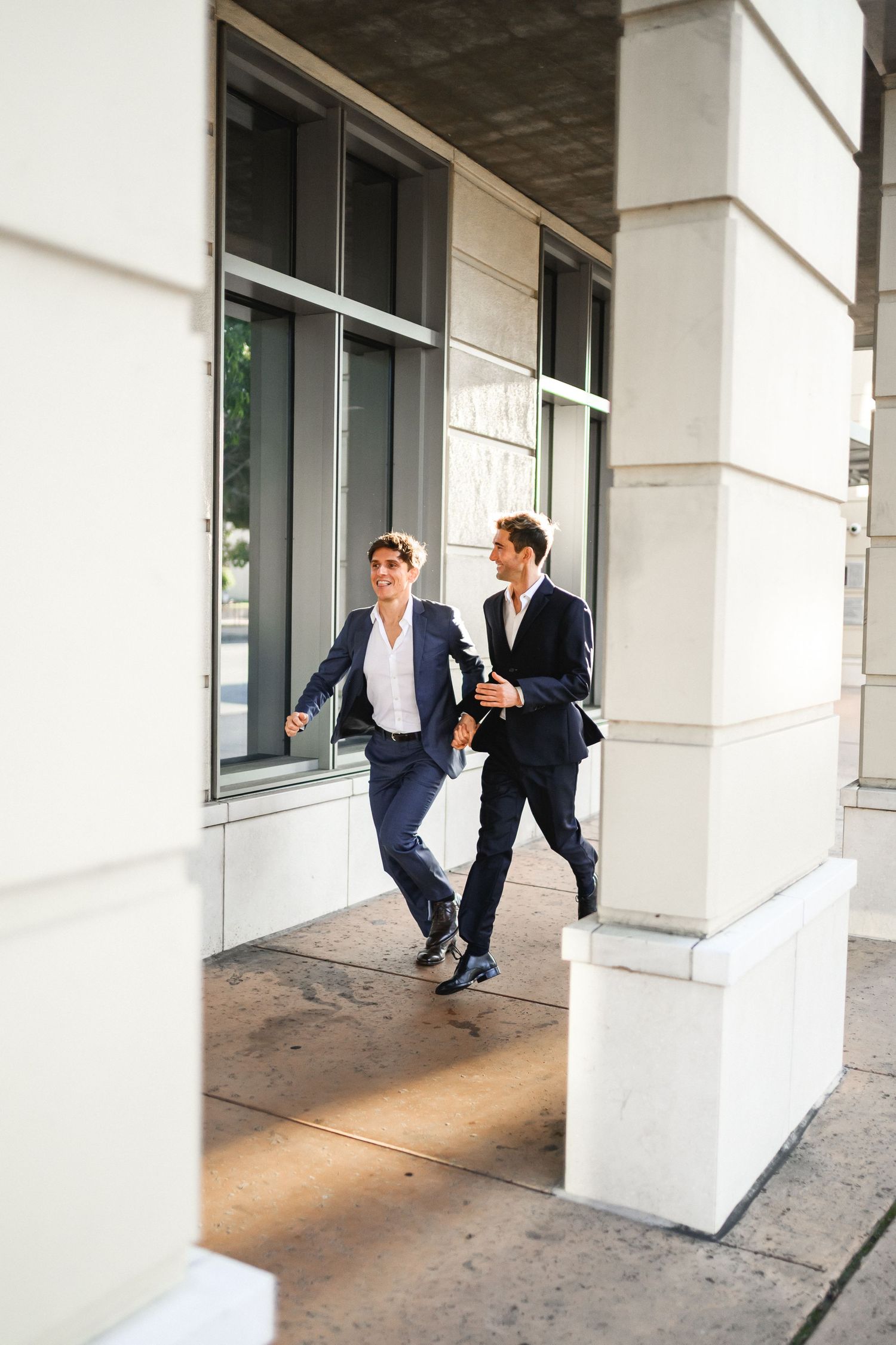 Two men in suits walking and laughing together outside a modern building in sunlight.