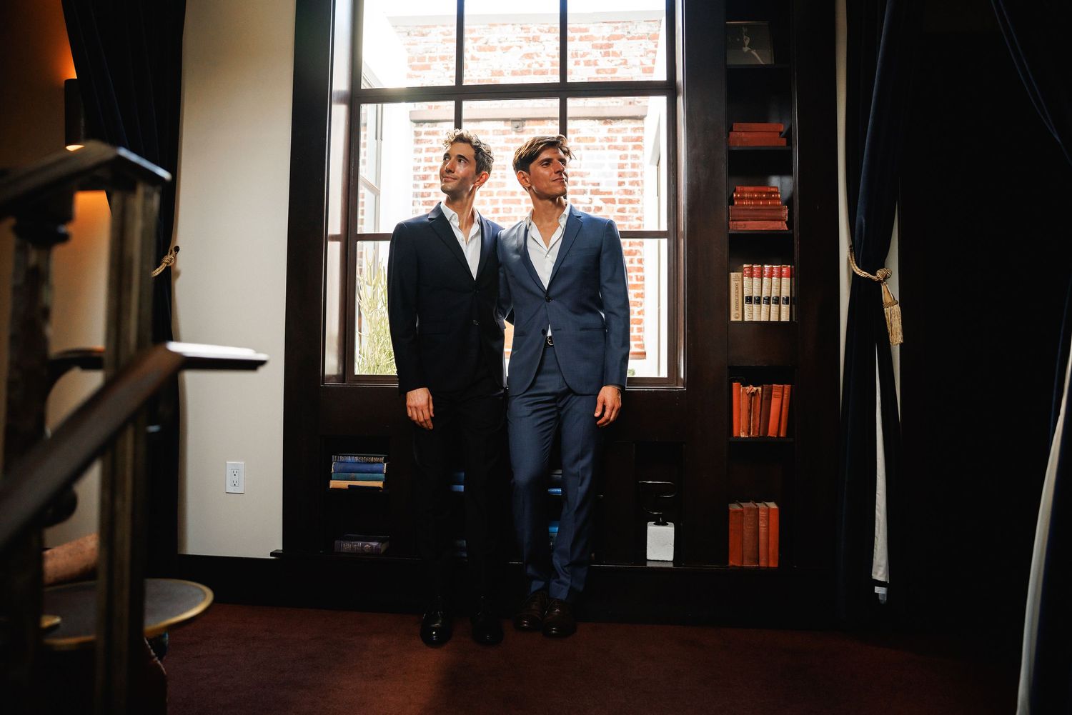 Two grooms in navy suits standing together in a library with large window backdrop.