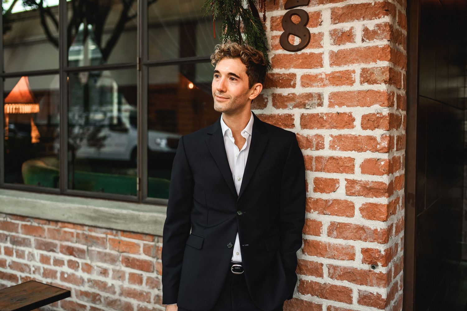 Stylish man in black blazer leaning against exposed brick wall outdoors.