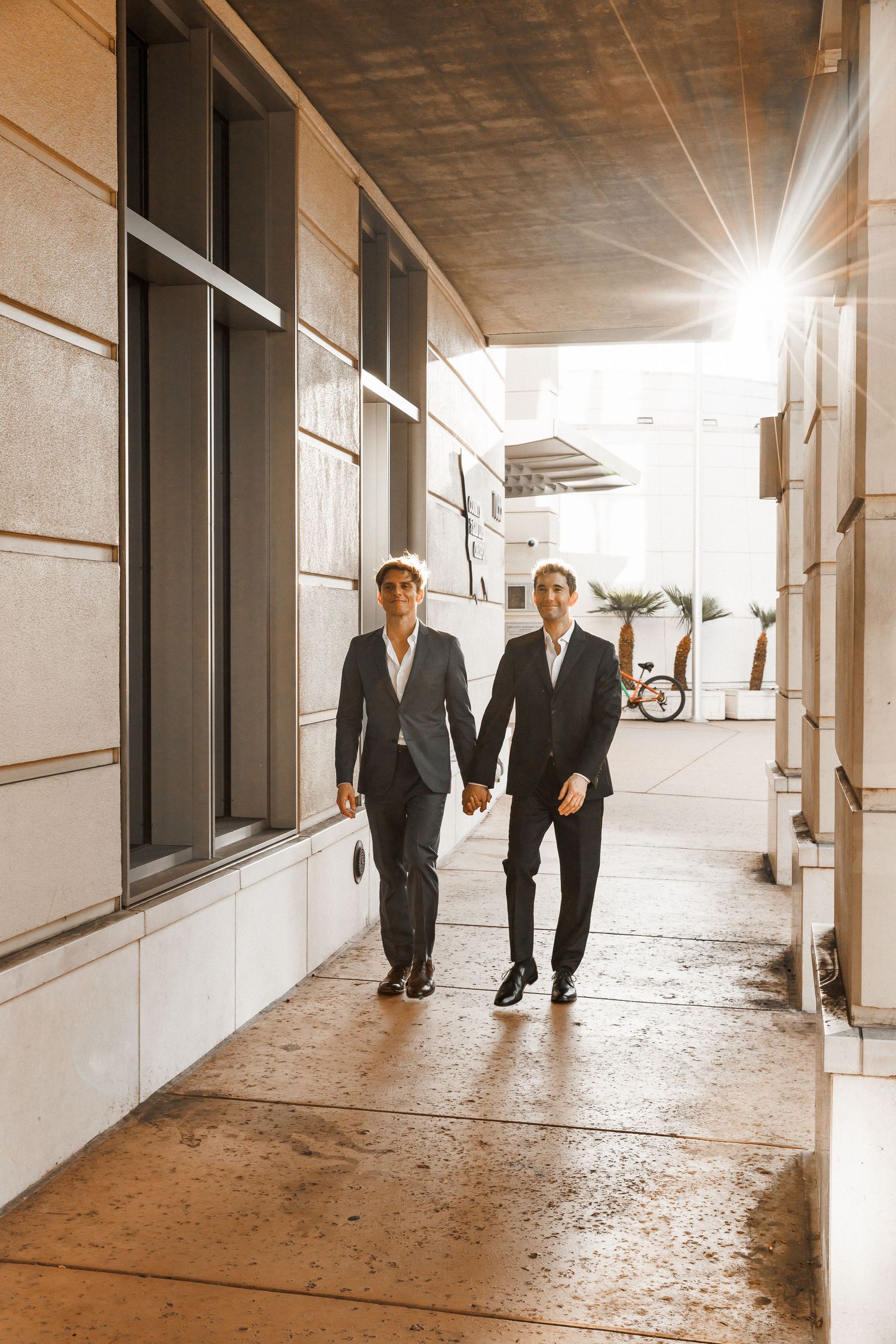 Two grooms holding hands walking through a sunlit urban corridor together.