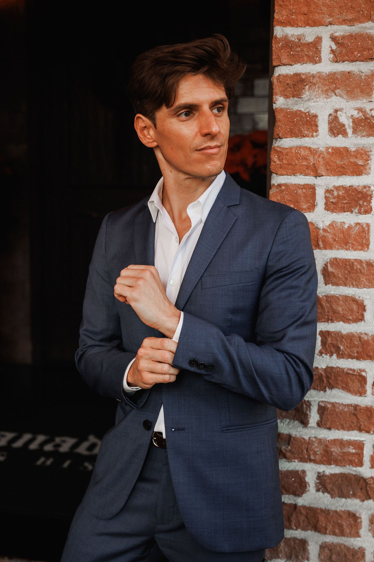 Groom in blue suit adjusting cufflinks while leaning against an exposed brick wall.