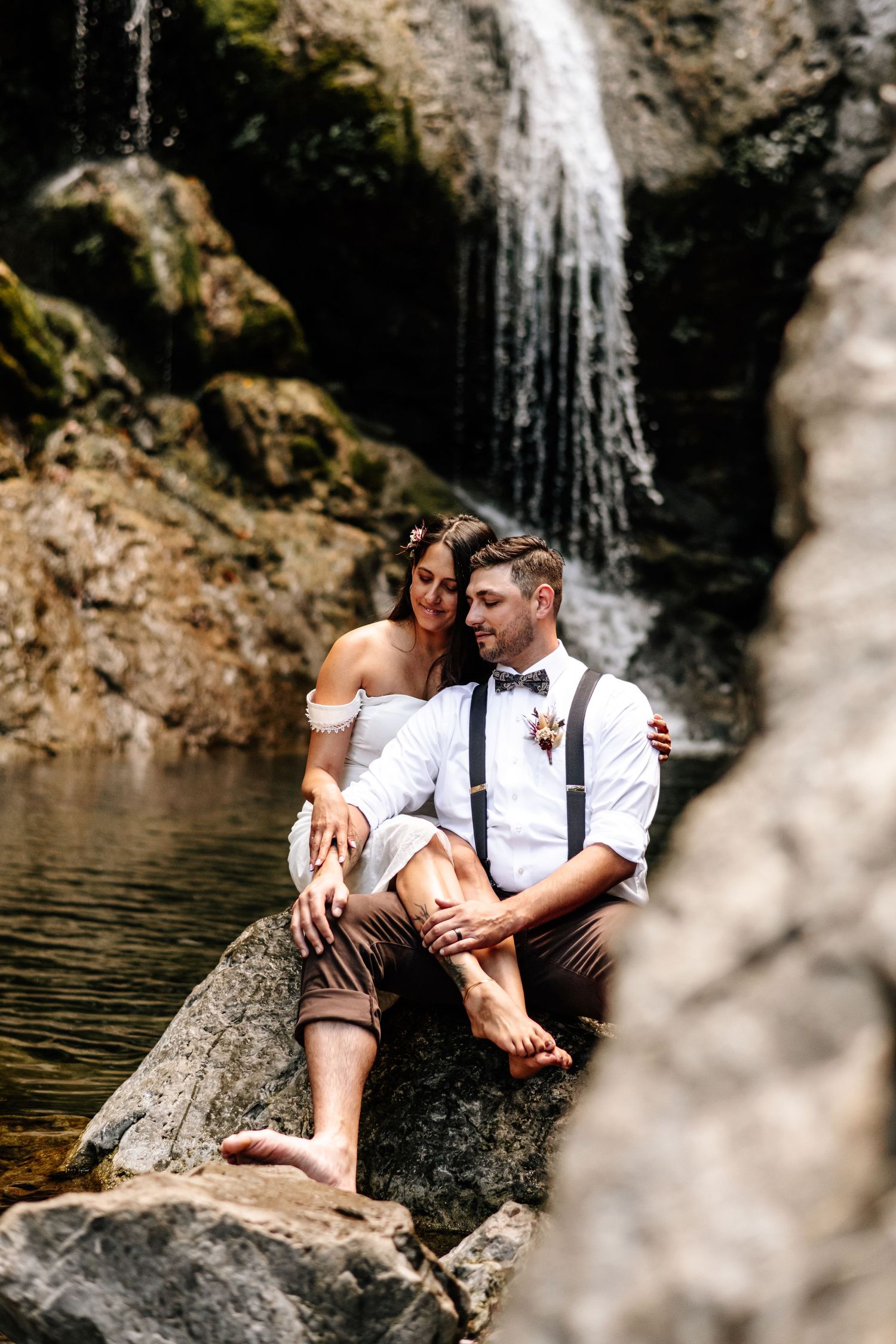 Bride and groom sitting barefoot on rocks embracing in front of scenic waterfall.