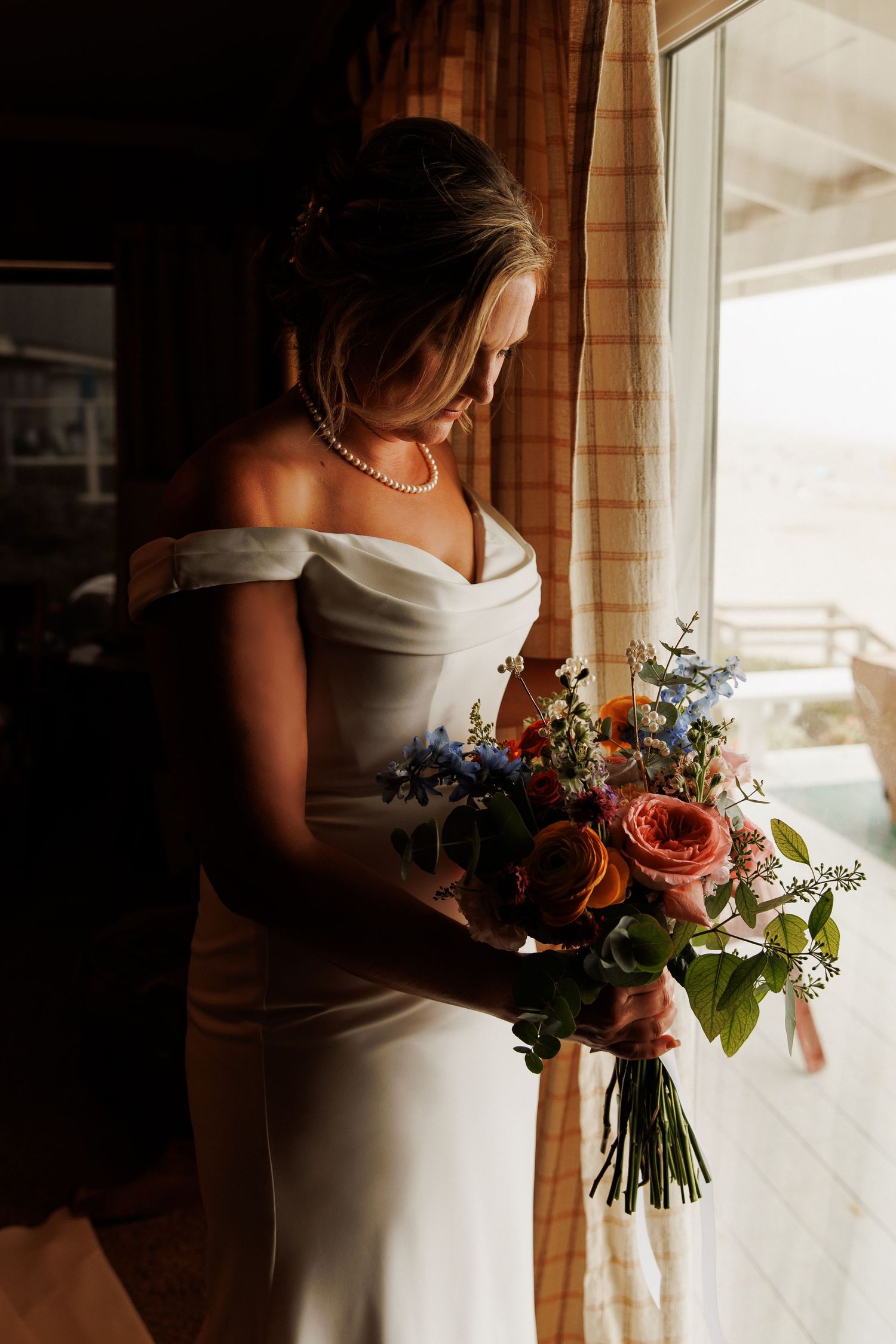 Bride in off-shoulder white gown holding colorful bouquet of roses and wildflowers by a sunlit window.