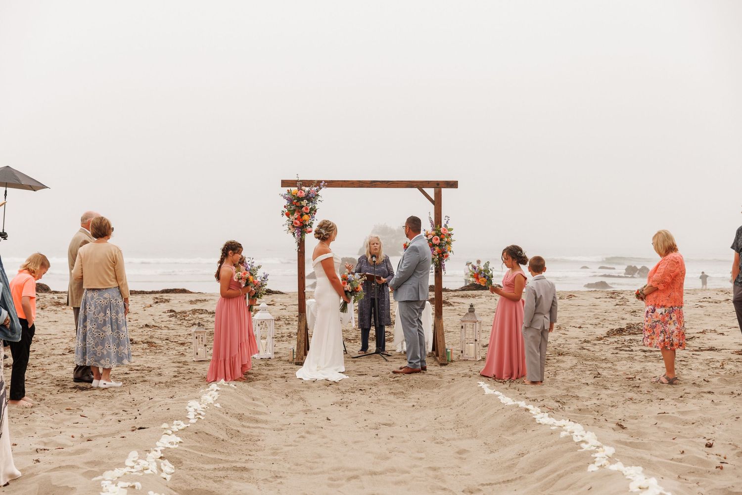 Intimate beach wedding ceremony at a wooden arch with small group of guests on a misty shoreline.