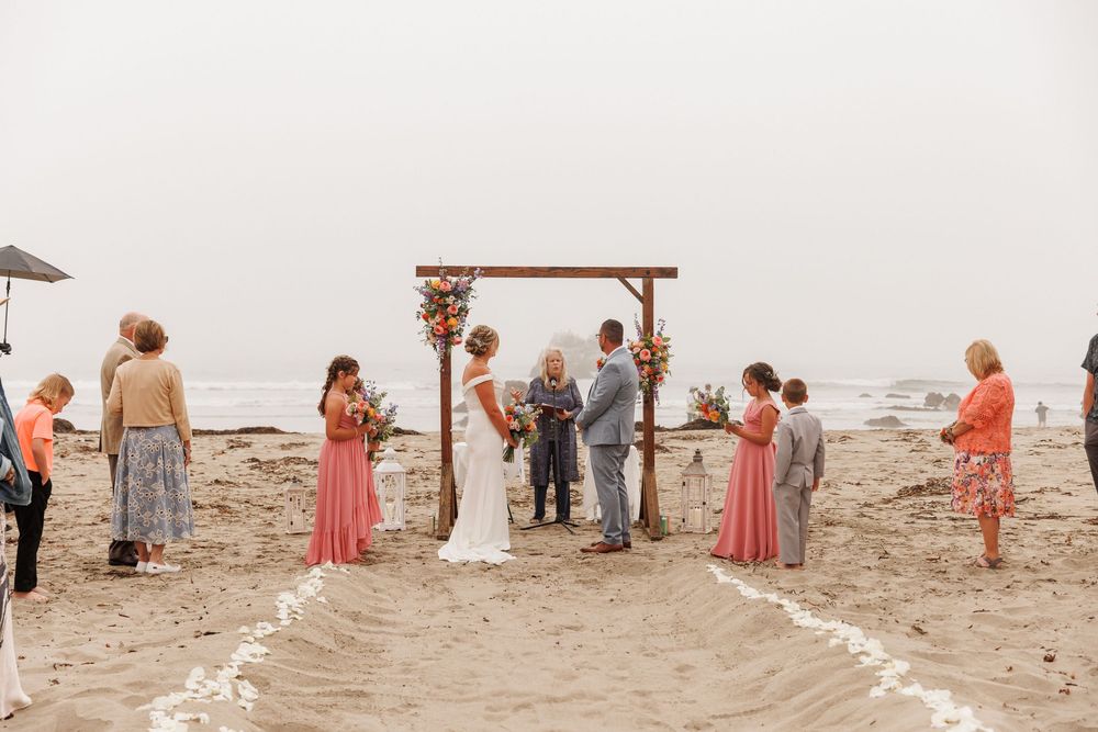 Intimate beach wedding ceremony at a wooden arch with small group of guests on a misty shoreline.