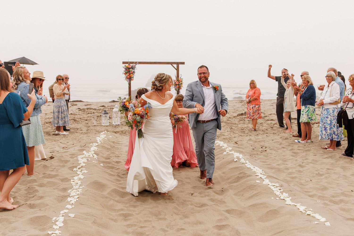 Joyful newlyweds walking back down sandy aisle after beach wedding ceremony with cheering guests.