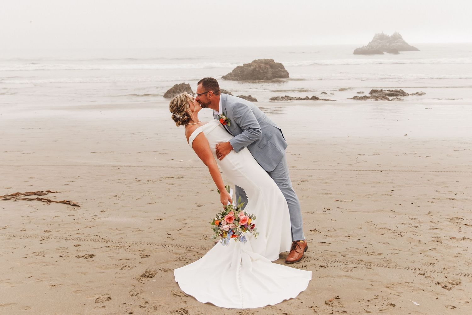 Groom dipping bride on a foggy beach, bride holding vibrant bouquet with rocky coast in background.