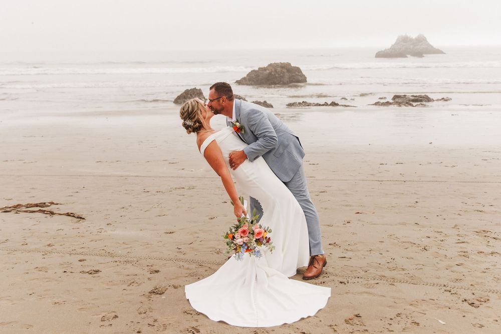 Groom dipping bride on a foggy beach, bride holding vibrant bouquet with rocky coast in background.