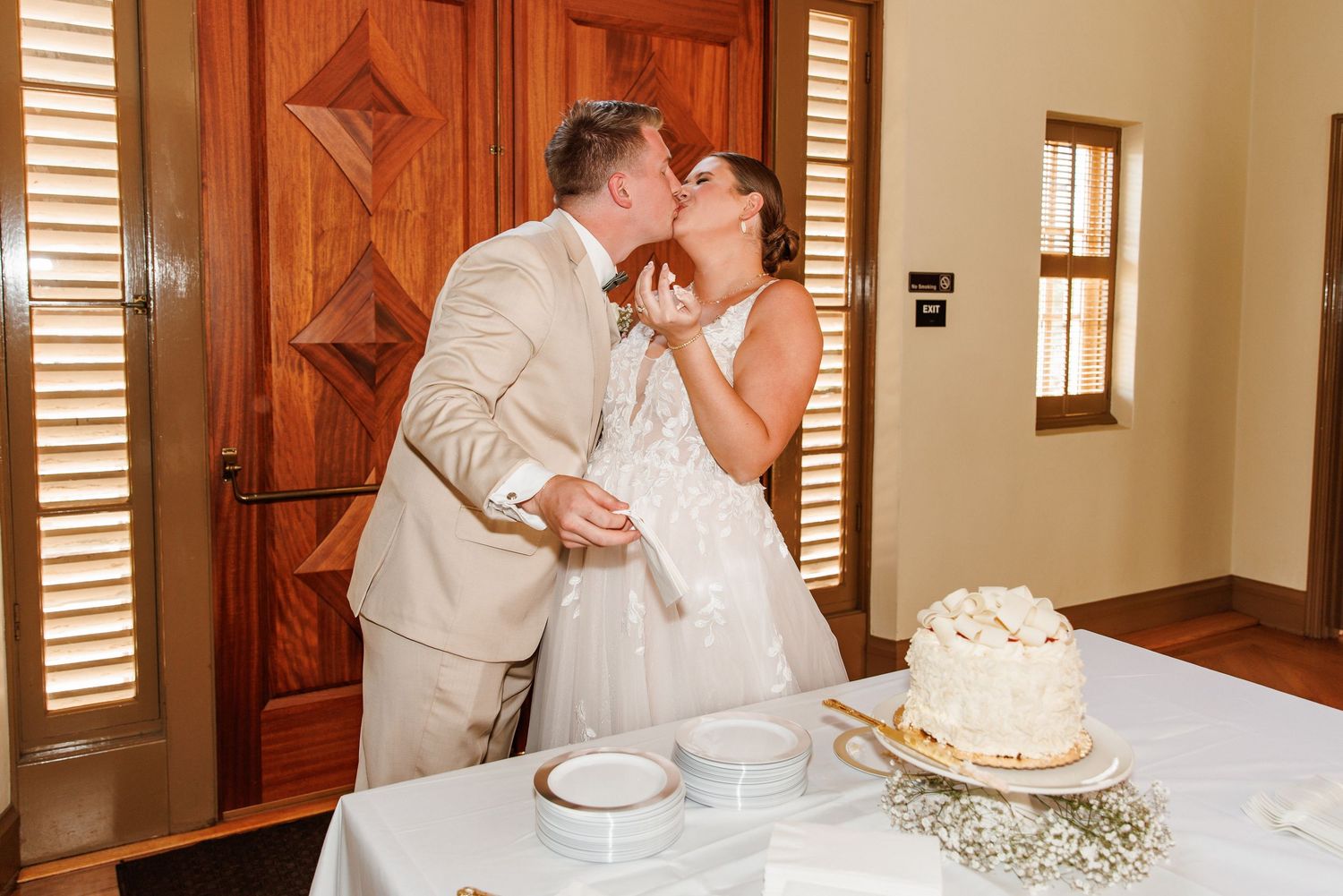 Newlyweds sharing a kiss while cutting their white wedding cake at reception.